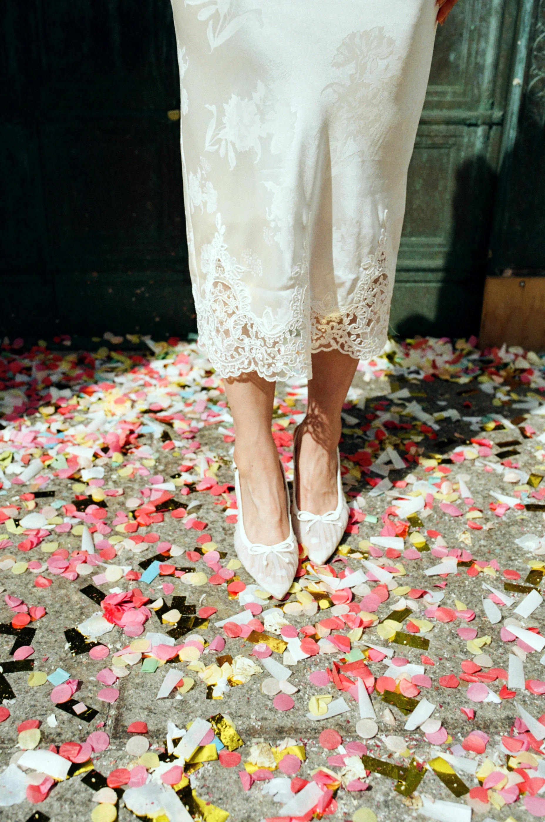 A woman standing on a ground covered with colorful confetti, wearing a white lace dress and white shoes.