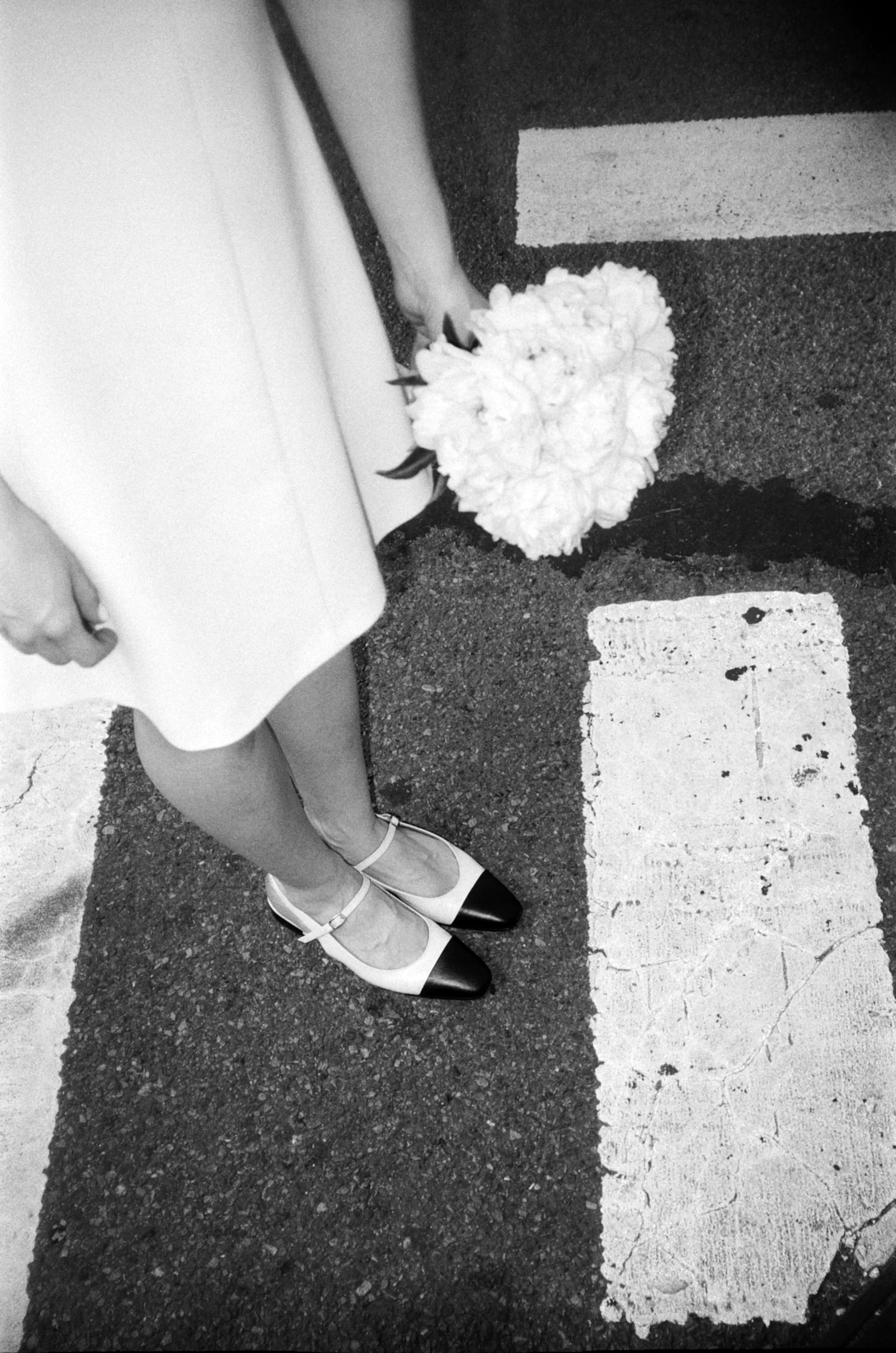 A person in a dress and heels holds a bouquet of white flowers, standing on a ground with painted parking lines.