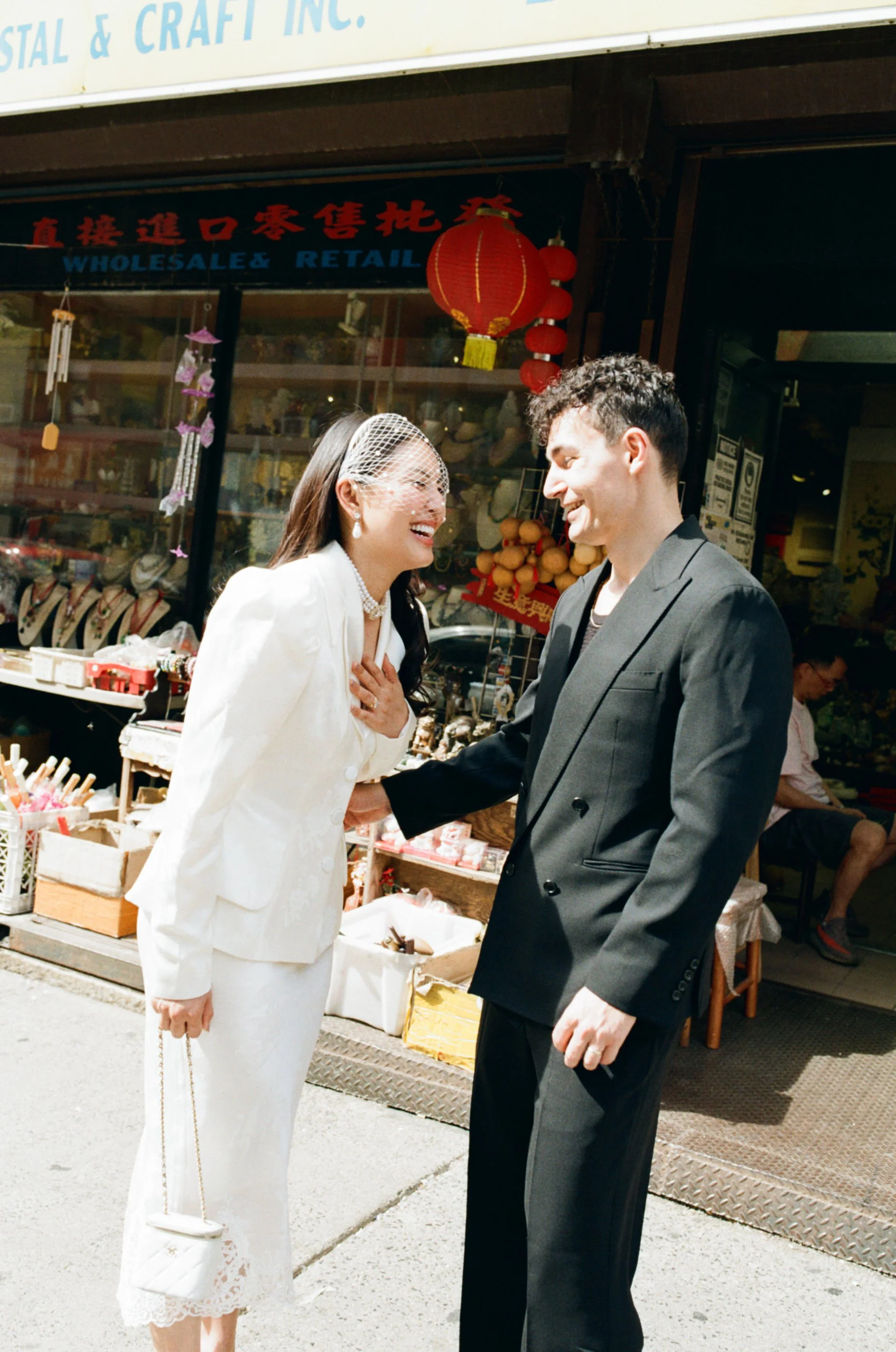 A man and a woman, dressed in formal attire, are smiling and shaking hands on a sidewalk outside a shop with red lanterns hanging above. The woman is wearing a white suit with lace trim and a birdcage veil, and the man is wearing a gray suit. They appear to be celebrating a special occasion.
