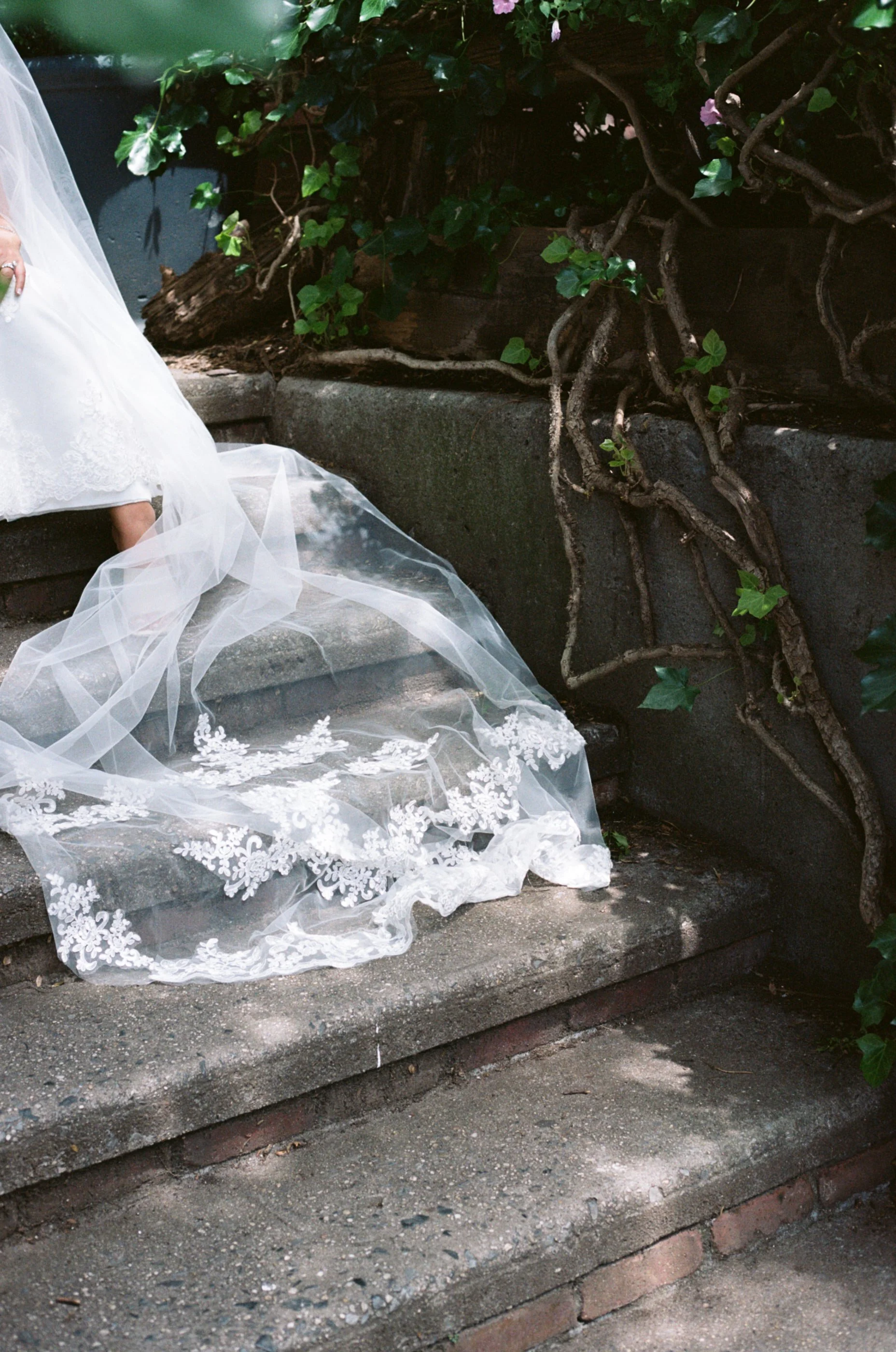 Part of a white wedding dress with lace and tulle fabric, hanging over concrete steps, with greenery and pink flowers in the background.