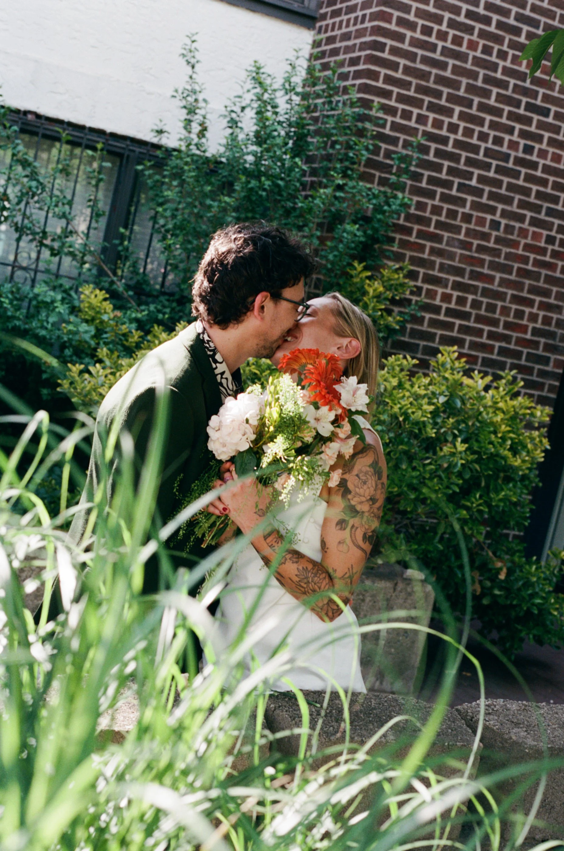 A couple sharing a kiss outdoors in a garden with greenery and a brick wall in the background. The woman has tattoos on her arms and holds a bouquet of flowers.