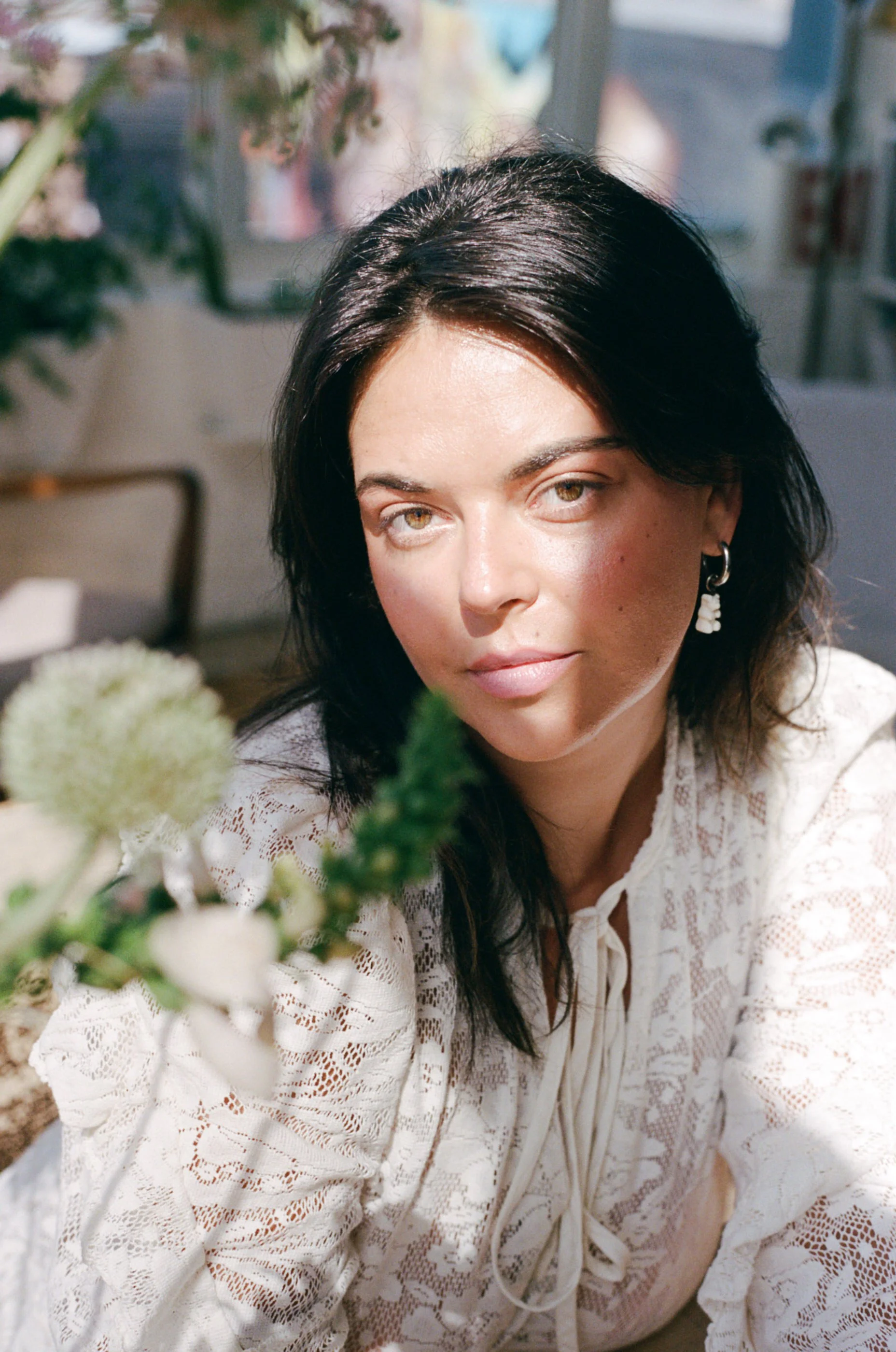 A woman with dark hair and brown eyes wearing a white lace top and earrings, surrounded by flowers, in bright natural light.