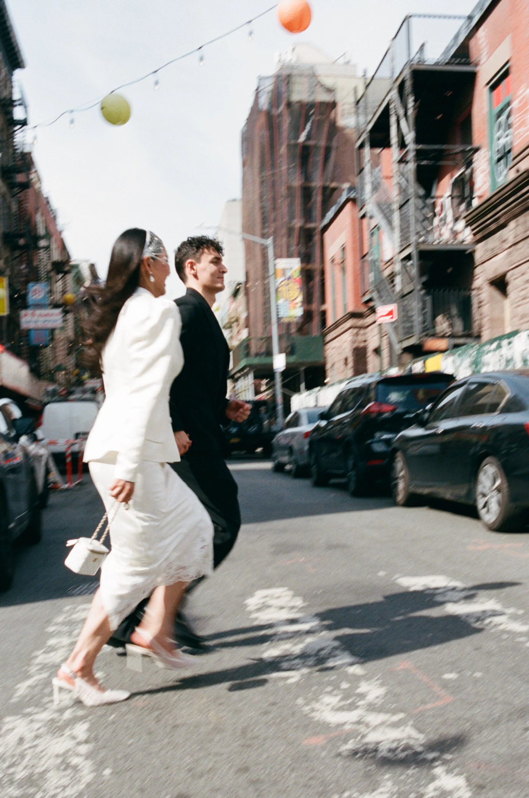 Two women and a man walking across a city street at a crosswalk, with cars parked along the curb and buildings in the background, under decorative string lights and paper lanterns hanging overhead.