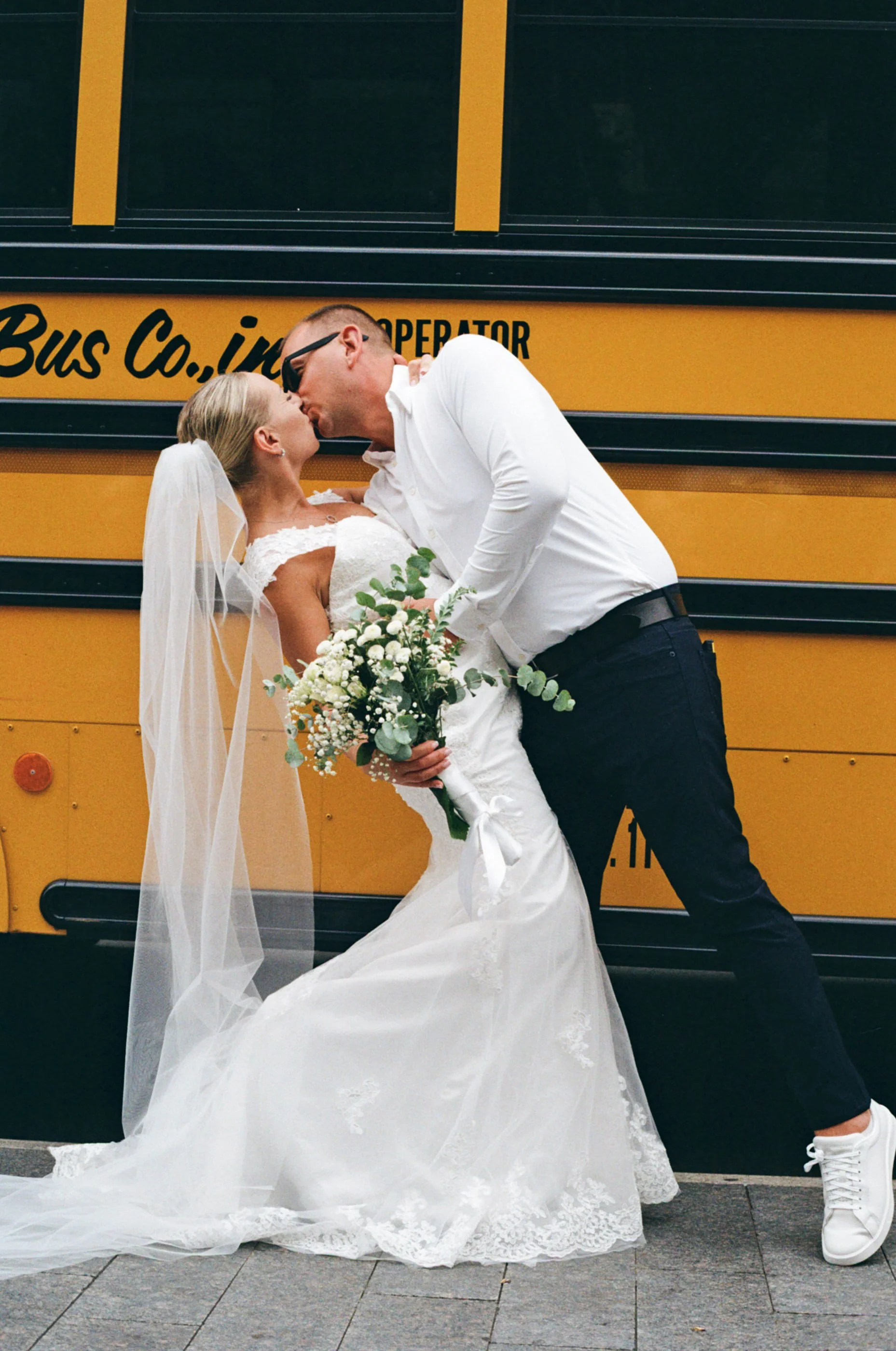 A bride and groom share a kiss in front of a yellow bus, with the bride holding a bouquet of white flowers and wearing a white wedding gown, while the groom is dressed in a white shirt and dark pants.