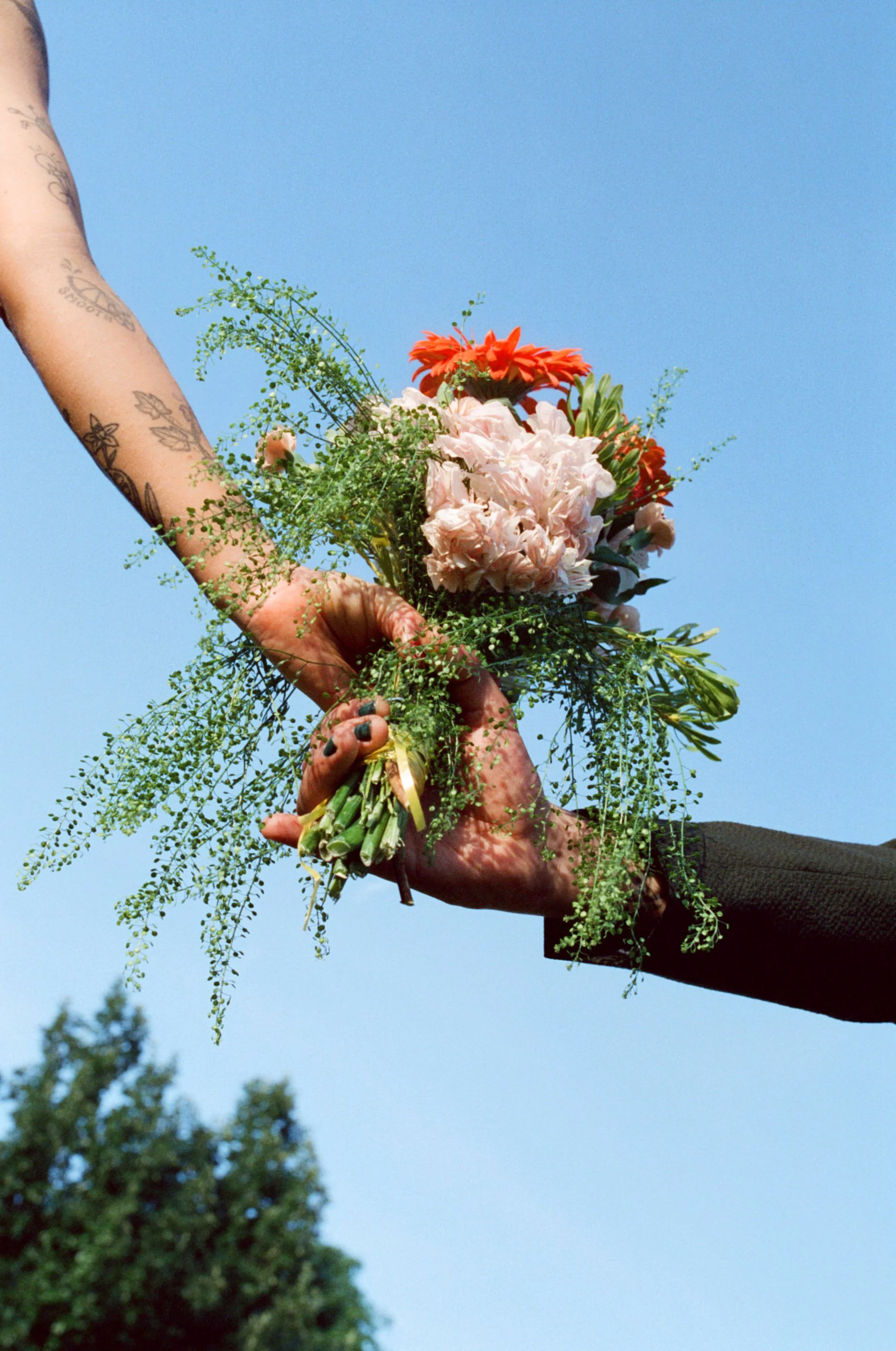 Two hands holding a bouquet of pink, orange, and white flowers against a blue sky.