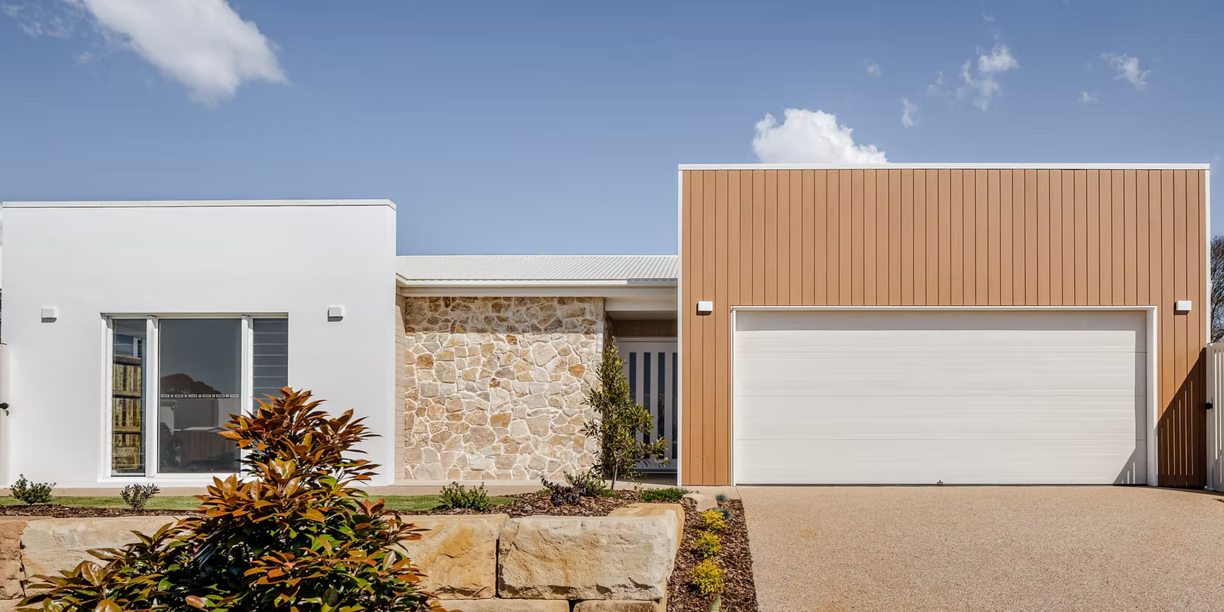 A modern house with a white facade on the left, a stone wall in the middle, and a reddish-brown wooden section with a garage door on the right. There are plants and small bushes in front, and a clear blue sky with a few clouds above.