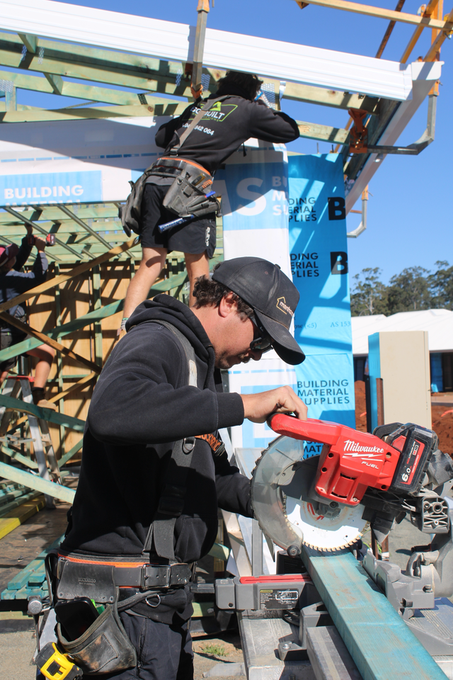 A construction worker operating a red Milwaukee circular saw cutting a piece of blue material on a construction site. Another worker is working on the structure above.