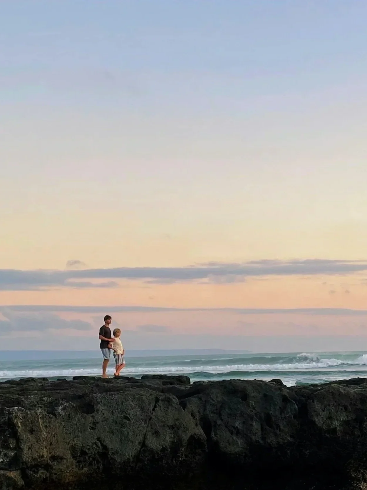 A man and a young girl standing on rocks near the ocean during sunset, with the sky painted in soft pastel colors and gentle waves in the background.