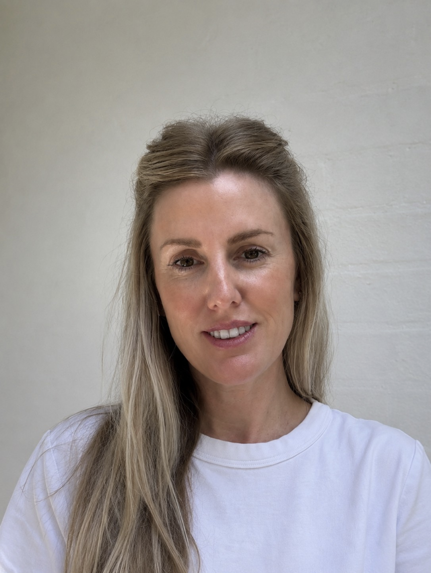 A woman with long blonde hair in a loose style, wearing a white t-shirt, smiling slightly, standing against a light-colored wall.