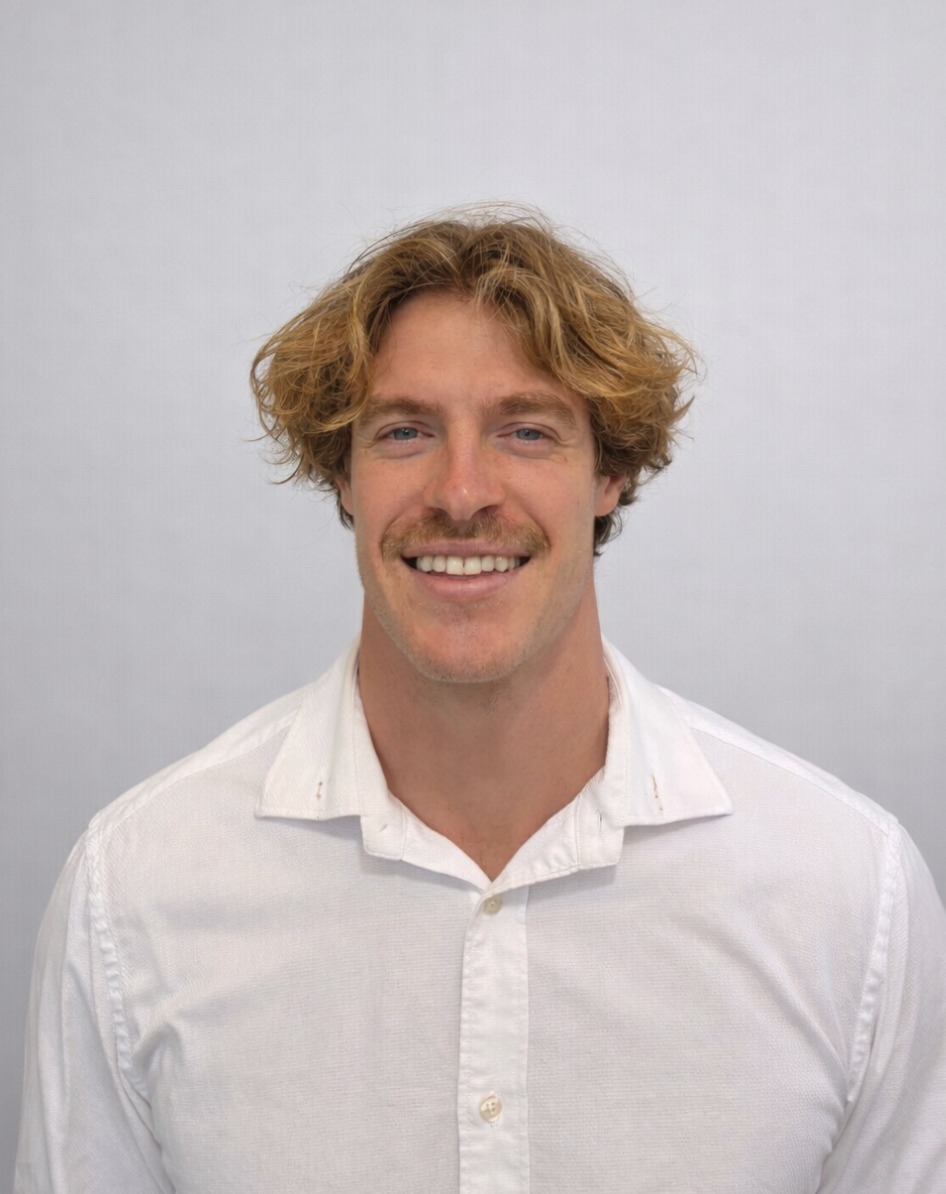 A smiling man with curly reddish-blonde hair, blue eyes, a light mustache, wearing a white button-up shirt, standing against a plain light gray background.