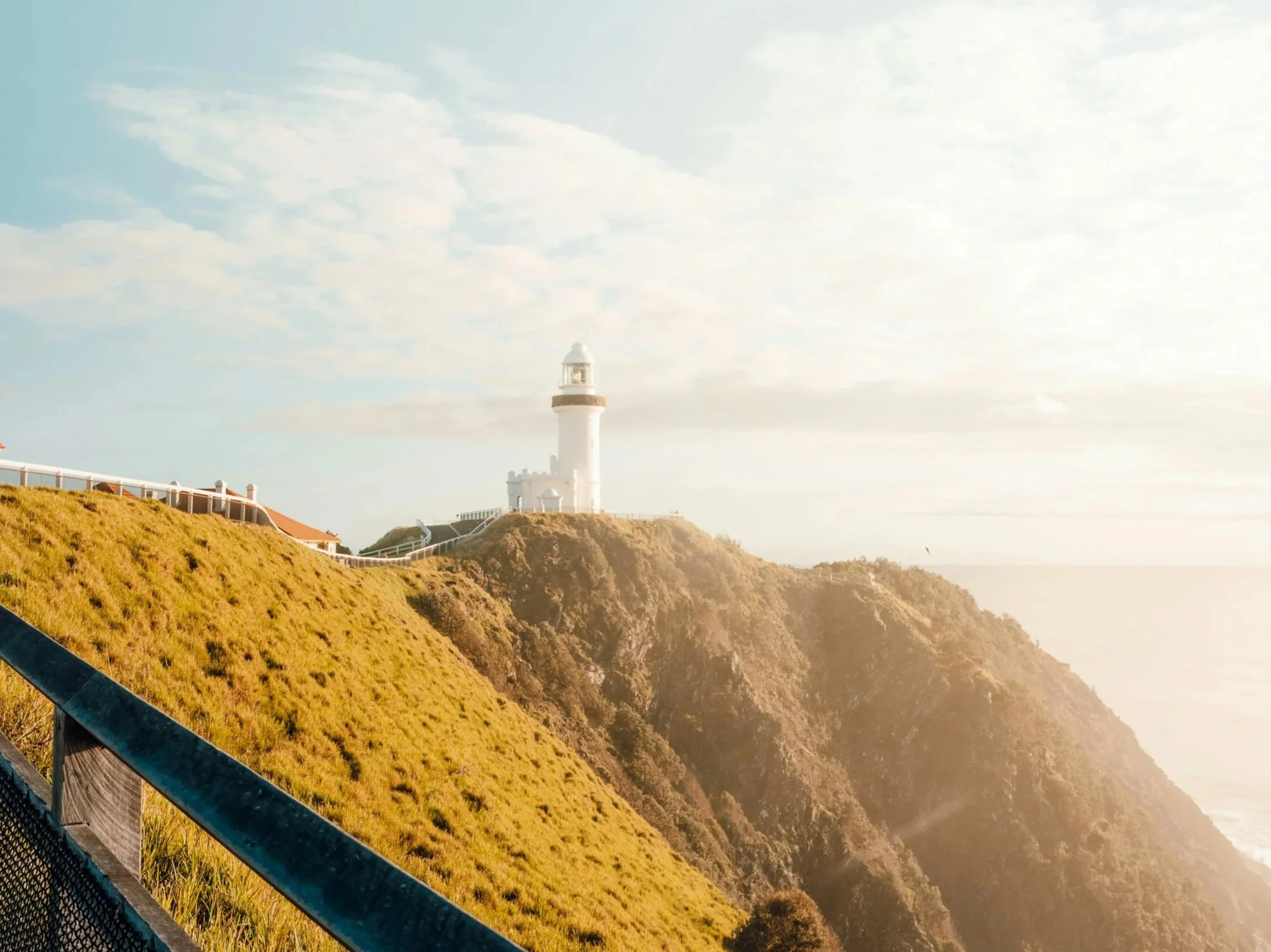 A lighthouse on a hilltop with a cloudy sky in the background, a walkway with a railing on the left side, and grassy terrain in the foreground.