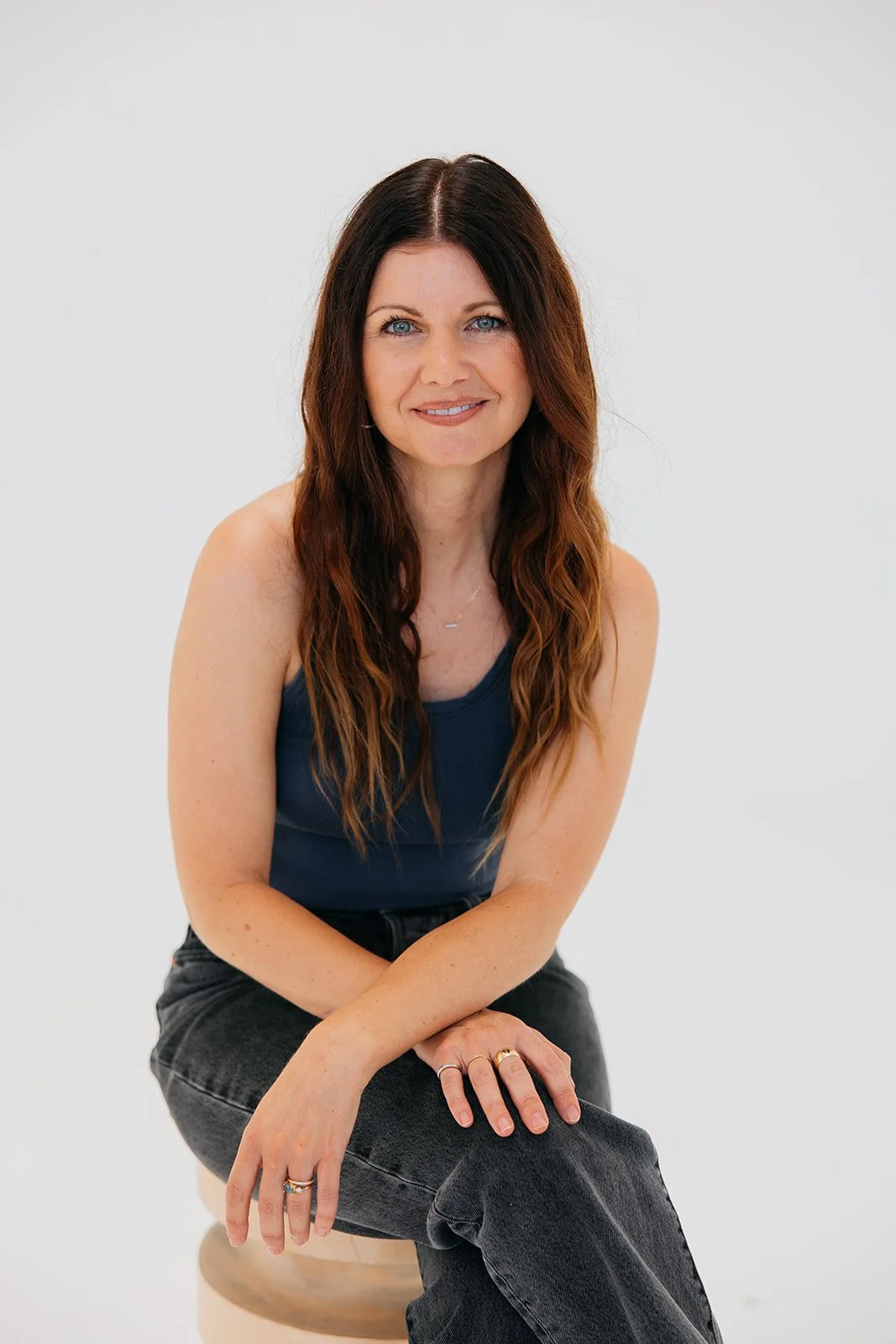 A woman with long, wavy brown hair and blue eyes sitting on a light-colored stool against a plain white background, wearing a navy sleeveless top and black jeans.