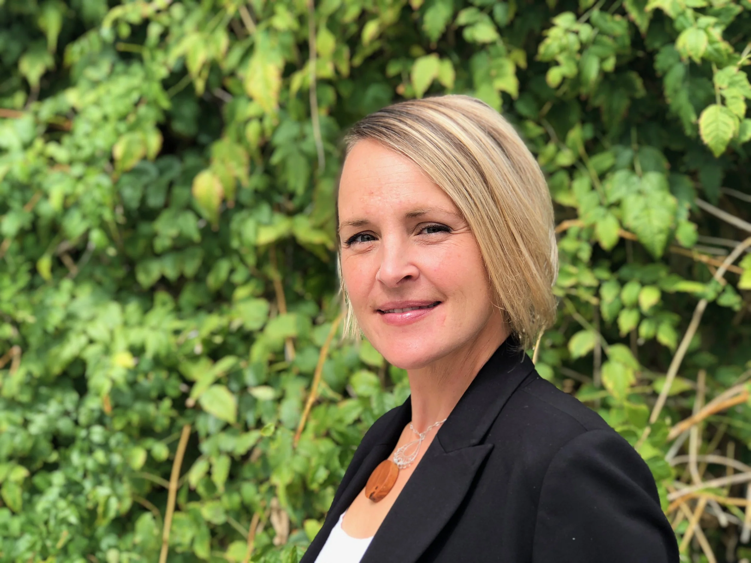A woman with blonde hair wearing a black blazer and white top, standing outdoors in front of green leafy bushes, smiling at the camera.