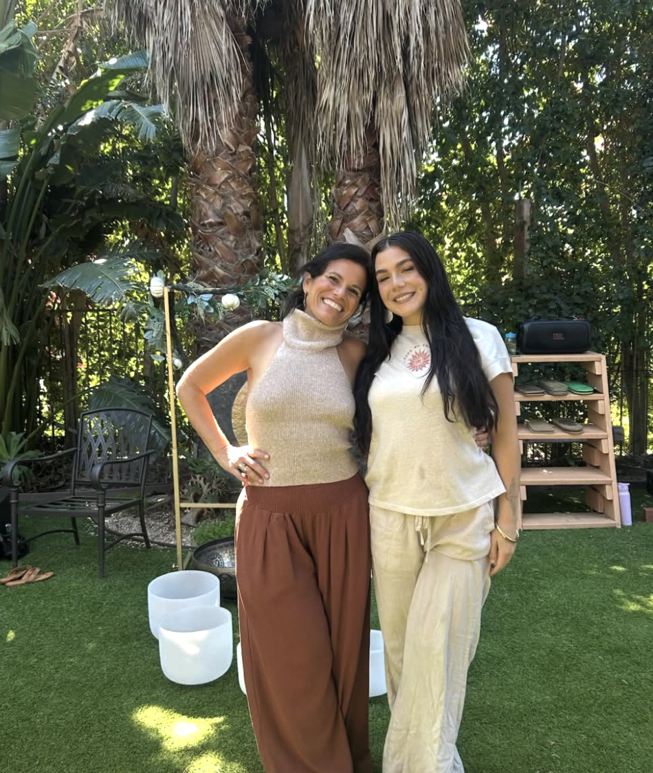 Two women standing close together outdoors in a lush garden. They are smiling, with one woman wearing a sleeveless beige turtleneck top and wide brown pants, and the other wearing a cream T-shirt and matching beige pants. Behind them are tall palm trees and various green foliage, with a wooden shelving unit, a black stereo, and garden chairs in the background.