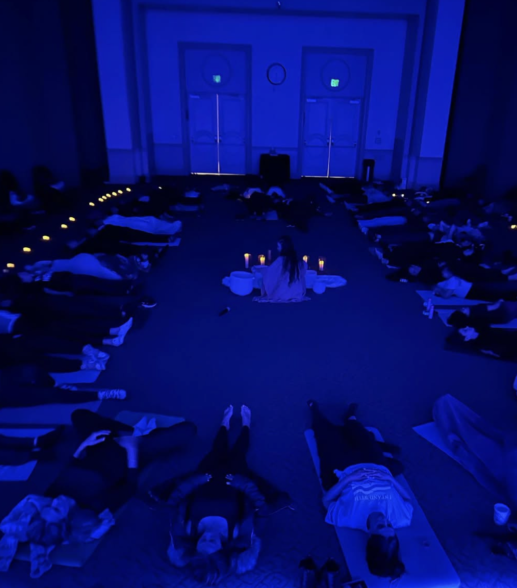 People lying on yoga mats in a dark room with blue lighting, arranged in a circle around a person sitting cross-legged, who appears to be leading a meditation or mindfulness session. Candles are placed in front of the instructor.