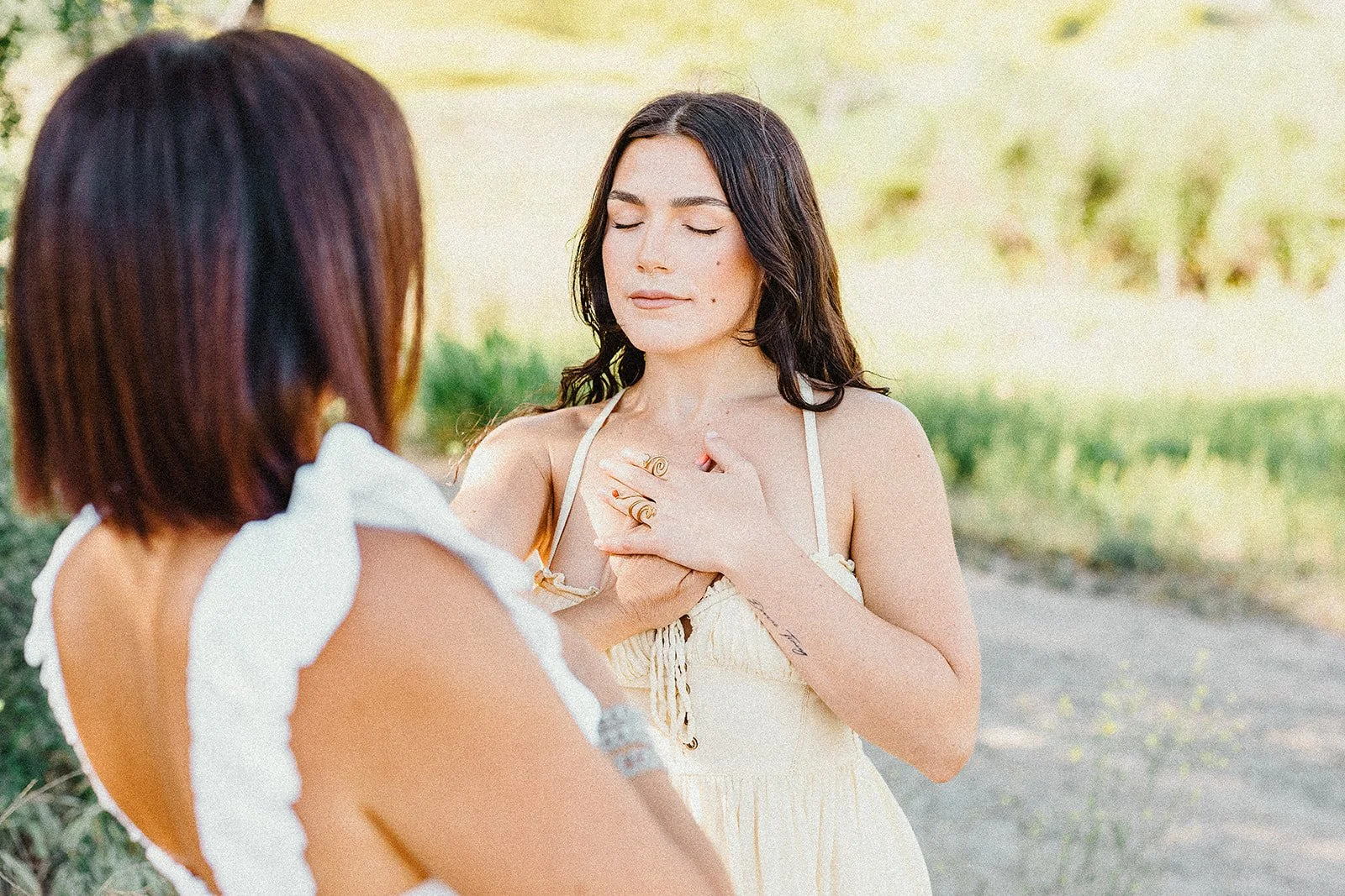 A woman with dark, curly hair and closed eyes places her hand on her chest during an outdoor ceremony, while another woman with short, reddish-brown hair faces her.