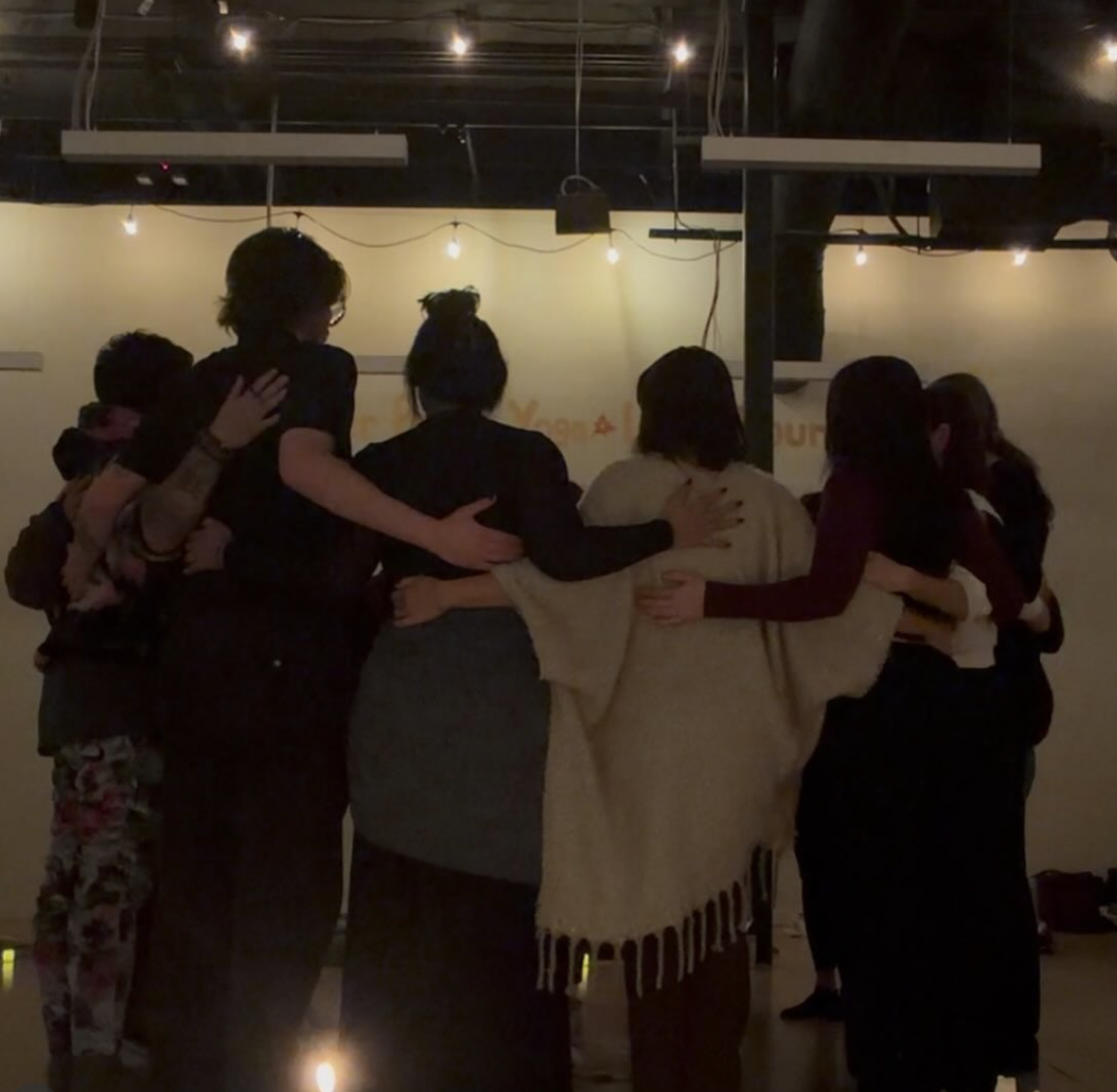 A group of people standing in a circle with their arms around each other's shoulders in a dimly lit setting.