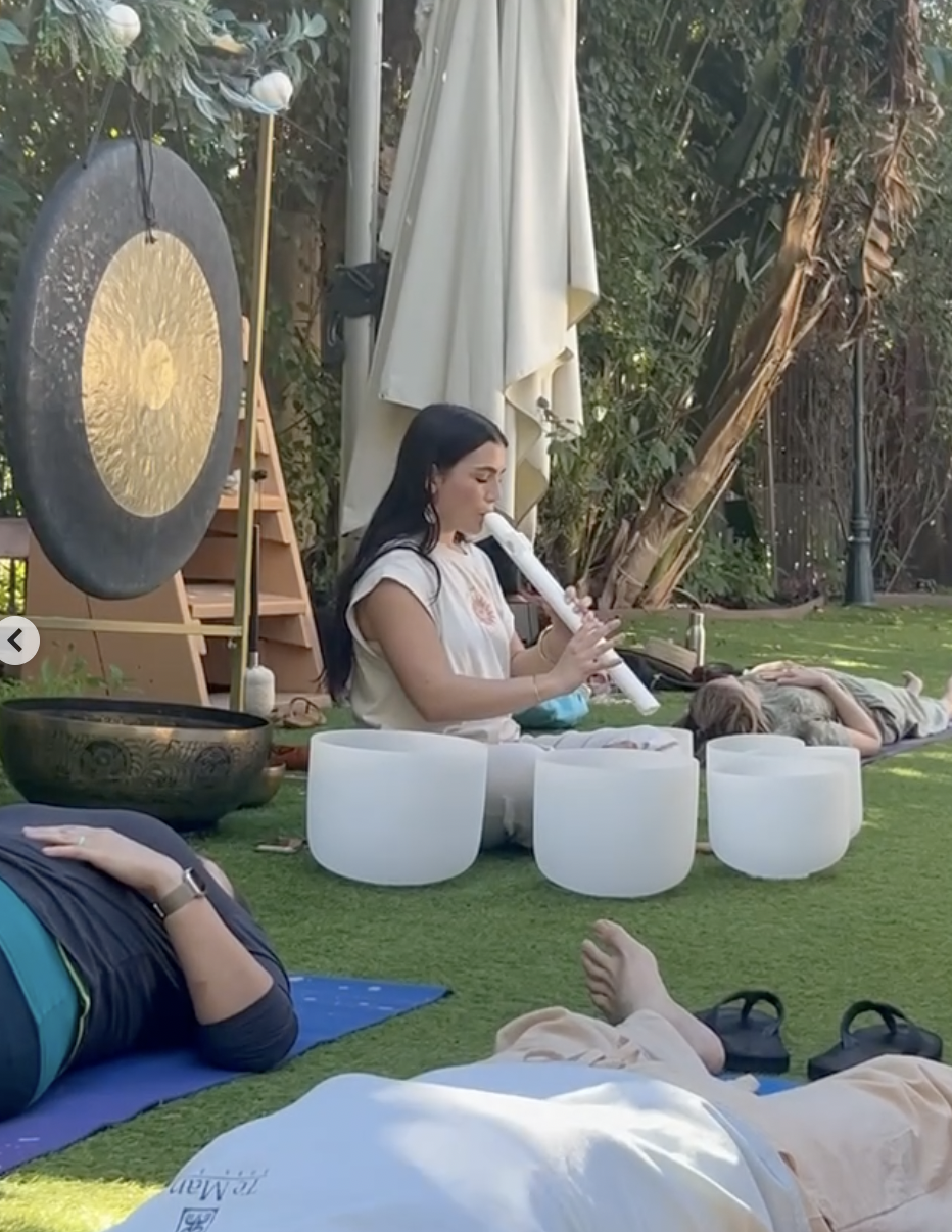 A woman playing a white flute while lying on the grass, surrounded by other people relaxing and practicing yoga outdoors with trees and a large gong in the background.