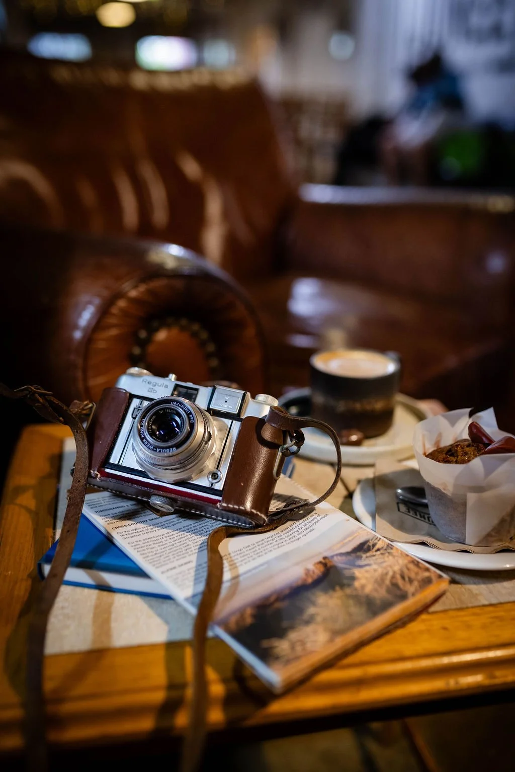 A vintage camera placed on an open book on a wooden table, with a cup of coffee, a bowl of food, and a magazine in the background