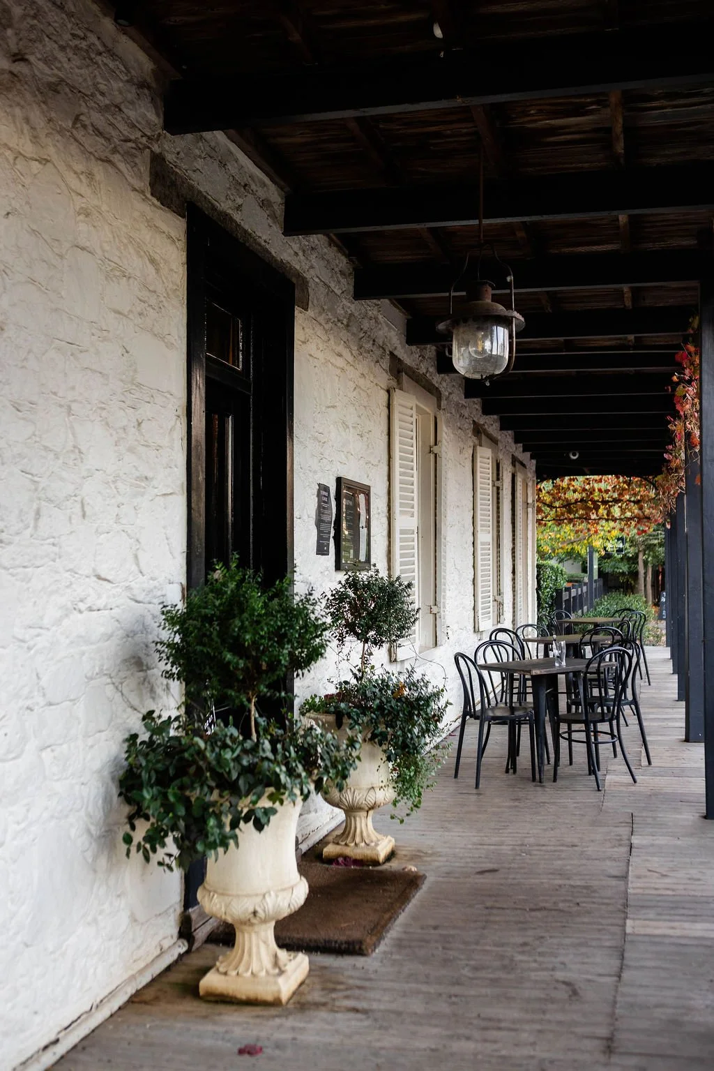 An outdoor patio with black chairs and wooden tables, white stone walls, potted plants, and hanging lamps beneath a dark wooden overhead roof.