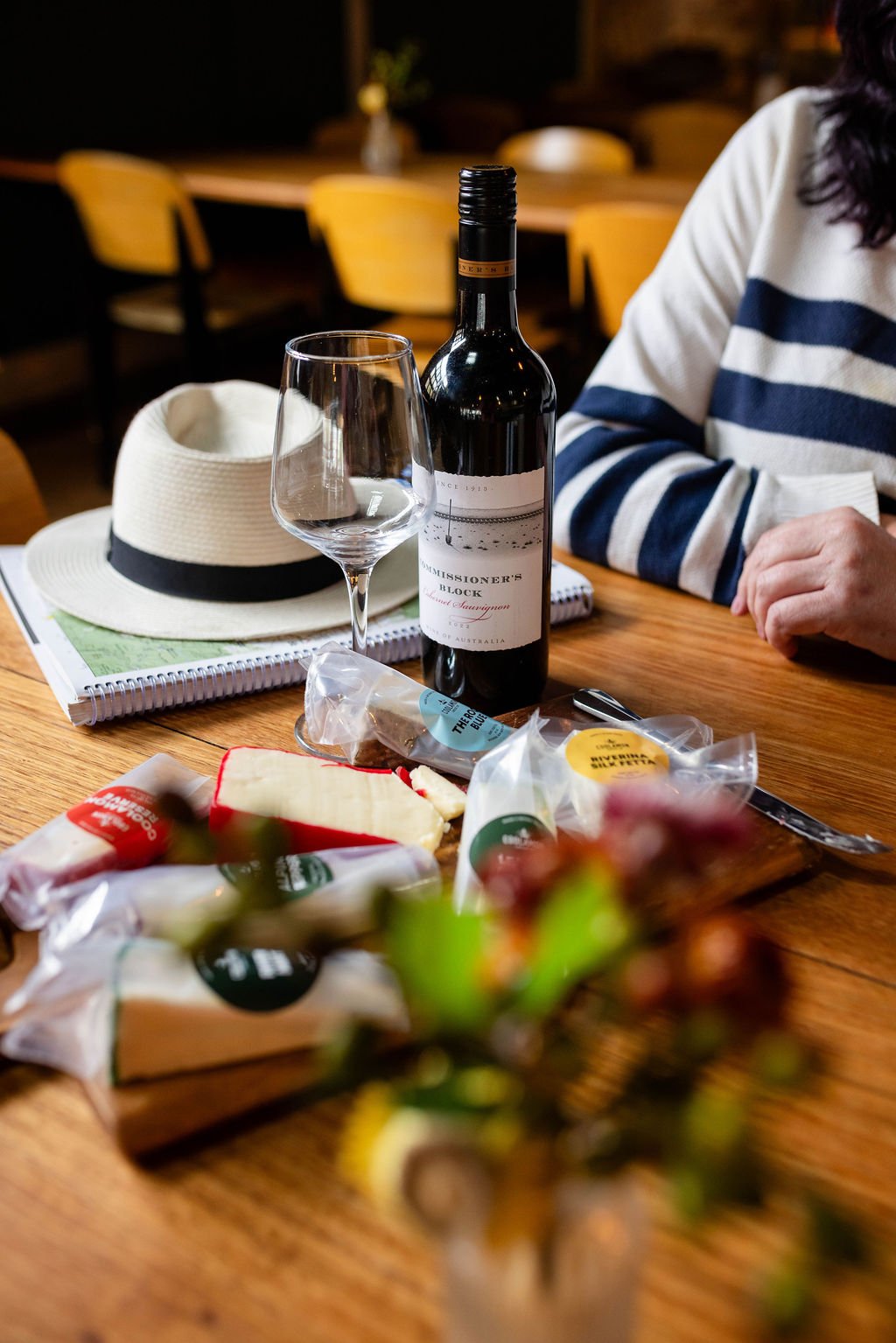 A bottle of red wine, an empty wine glass, a straw hat, and various cheeses on a wooden table. In the background, there are empty chairs and a person wearing a striped shirt. Some snacks are also on the table, with blurred flowers in the foreground.