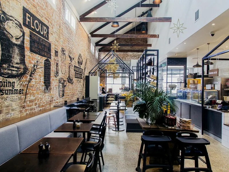 Interior of a cafe or bakery with black tables, chairs, and pastry display, brick wall with chalkboard art about flour, potted plants, and hanging star decorations.