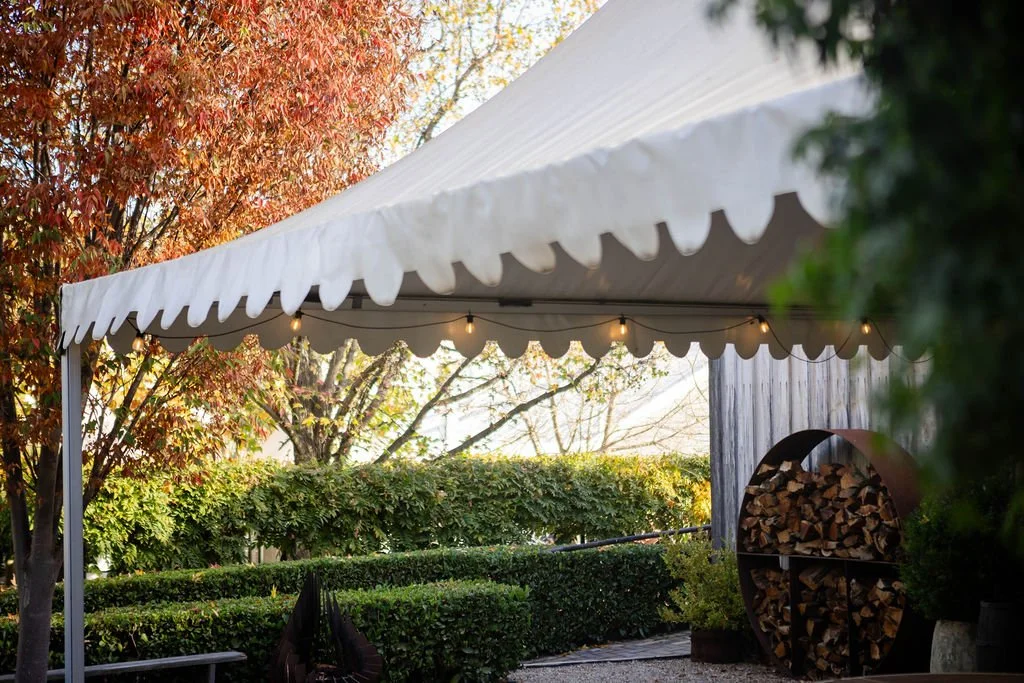 Outdoor scene with a white canopy tent, string lights, trimmed bushes, trees with autumn leaves, wood pile, and a garden area.