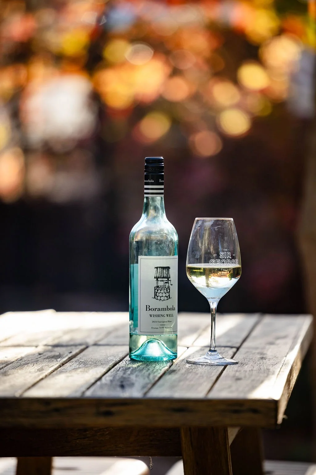 A bottle of Borambola wine and a filled wine glass on a wooden table outdoors during sunset, with blurred autumn trees in the background.