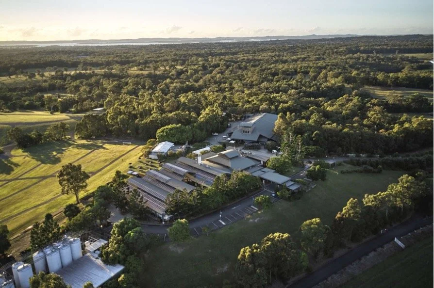 Aerial view of a farm with greenhouses, silos, and open fields surrounded by trees, with a forest in the background.
