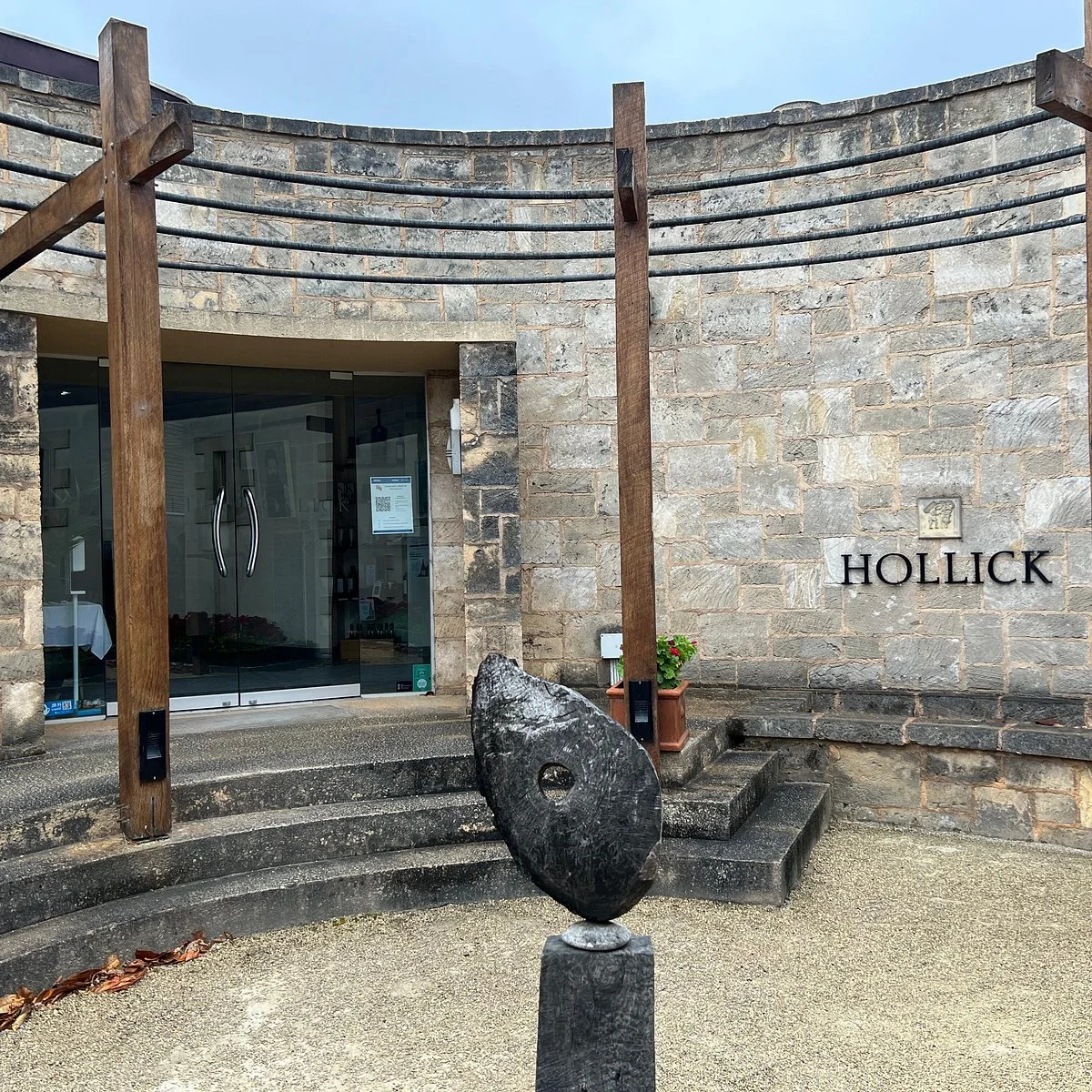 Exterior view of the Hollick winery with a stone building, a wooden arch framework, and a modern black stone sculpture in front of steps leading to the entrance.