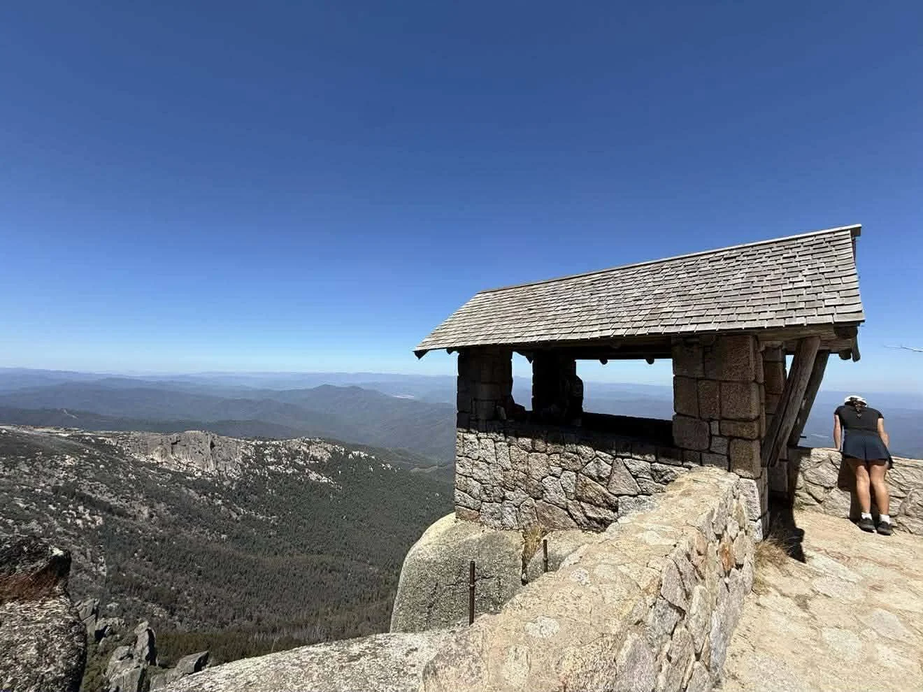 A small stone and wood shelter on a mountain overlook with a view of forested hills and mountains in the distance, under a clear blue sky.