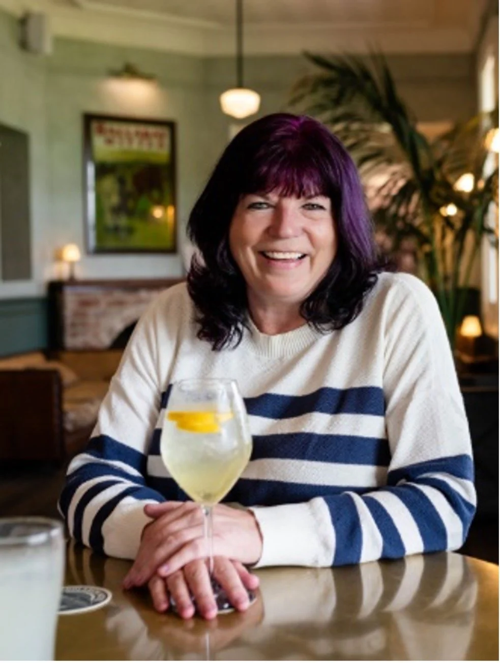 A woman with black and purple hair smiling at the camera, seated at a table with a glass of lemon water, in a cozy, well-lit restaurant or cafe with green walls and warm lighting.