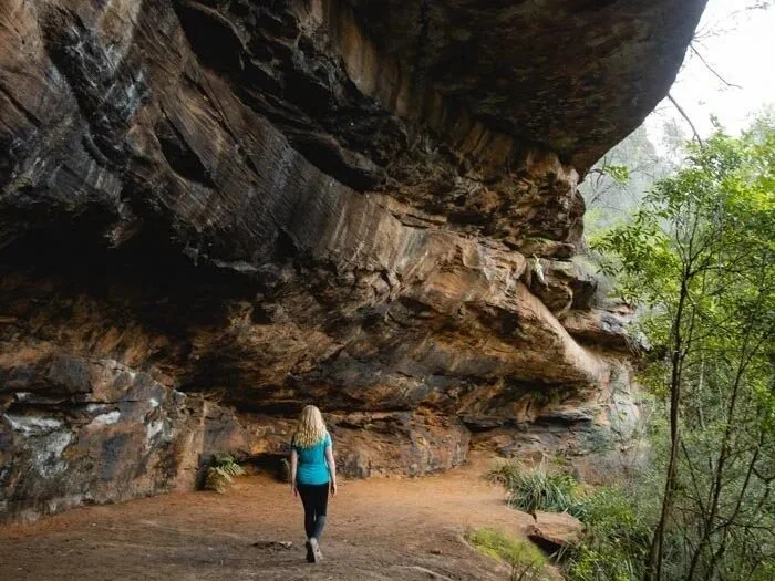 A woman with blond hair wearing a turquoise top and black pants walking on a dirt trail underneath a large overhanging rock formation in a forest.