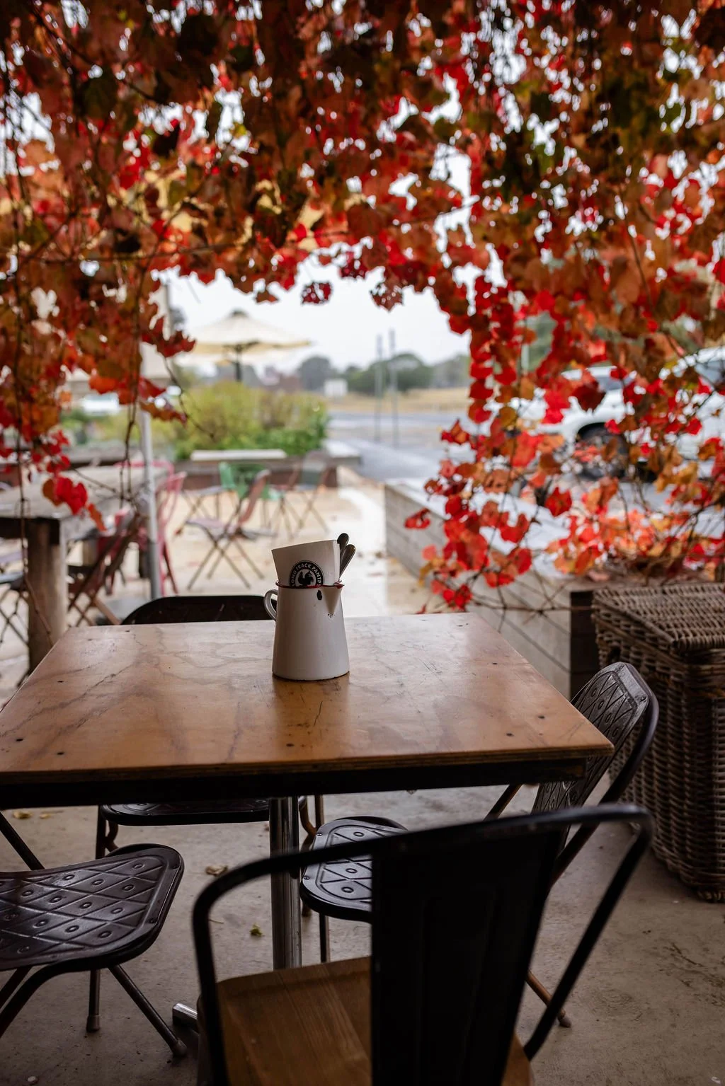 Empty outdoor cafe table with a white pitcher holding napkins and utensils, framed by autumnal red and orange leaves in the background.