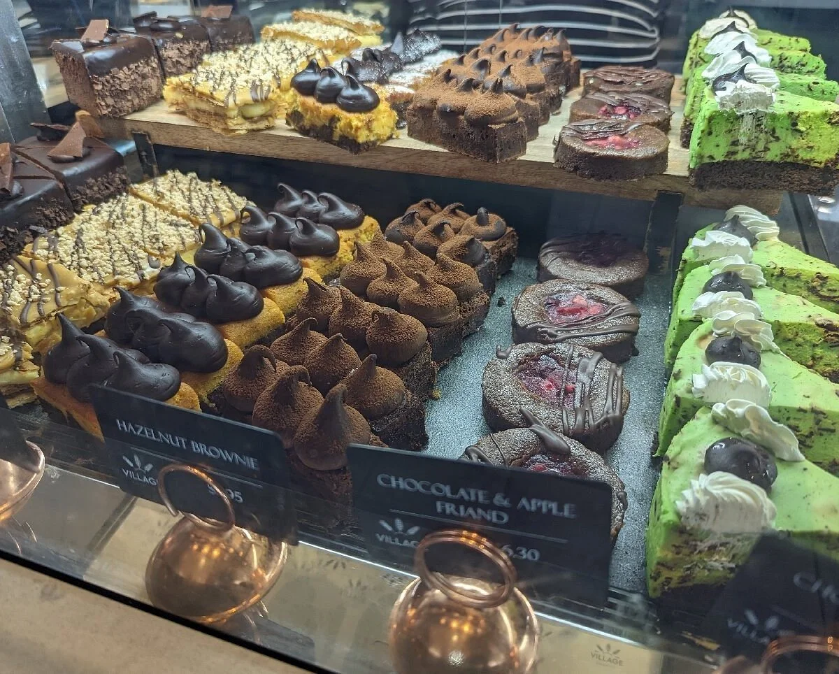 Assorted slices of various cakes and pastries displayed in a glass case, including hazelnut brownies, chocolate and apple friand, and green tea cakes topped with whipped cream and cherries.