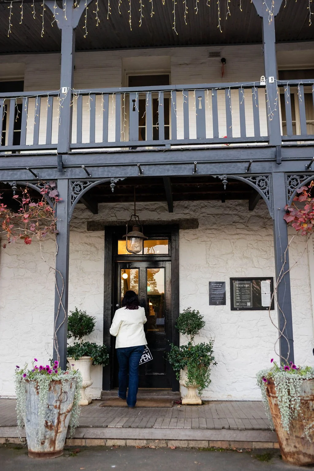 A person with dark hair, wearing a white jacket and blue jeans, stands in front of a black door with a glass panel, outside a restaurant or café. The building has white brick walls, with two large potted plants on either side of the door. There are two signs on the wall to the right of the door, and a hanging lantern above the entrance. The area is decorated with string lights and there are two large decorative flower pots with plants and flowers in the foreground.