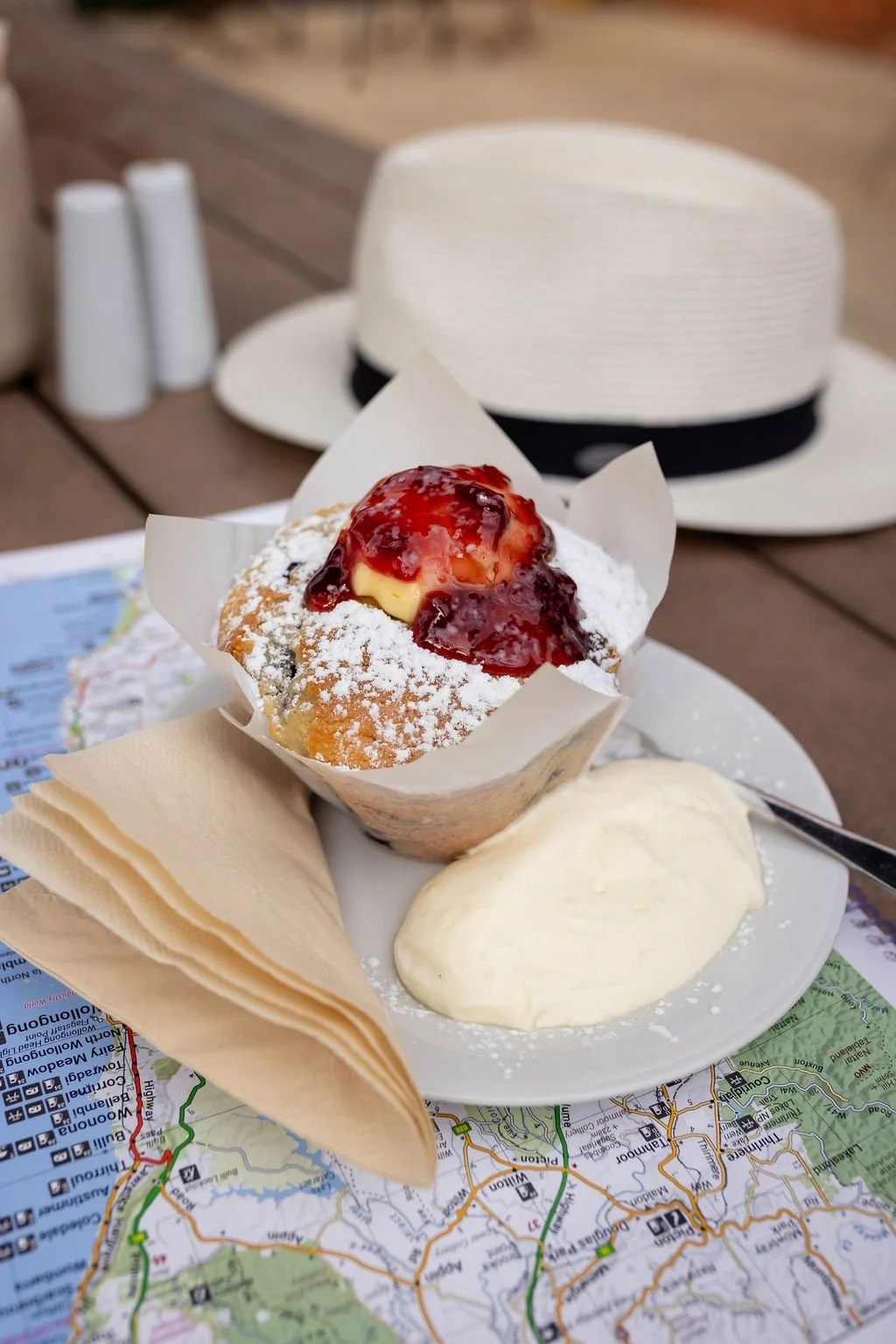 A cupcake with powdered sugar, topped with berry sauce and a scoop of butter or cream, served on a plate with a spoon, alongside a sheet of parchment paper, on top of a map. In the background, a white hat with a black band is visible, along with salt and pepper shakers.