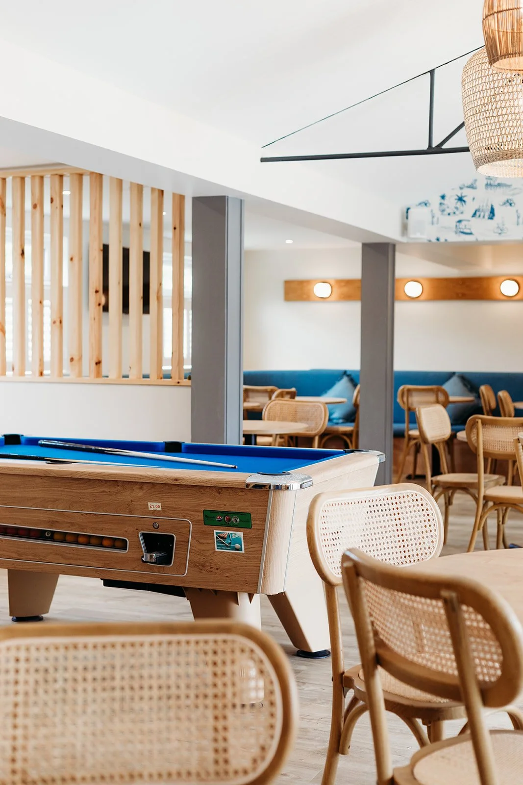 A recreational room with a wooden pool table, beige wicker chairs, and a blue cushioned seating area in the background. The decor includes modern lighting and wooden accents.