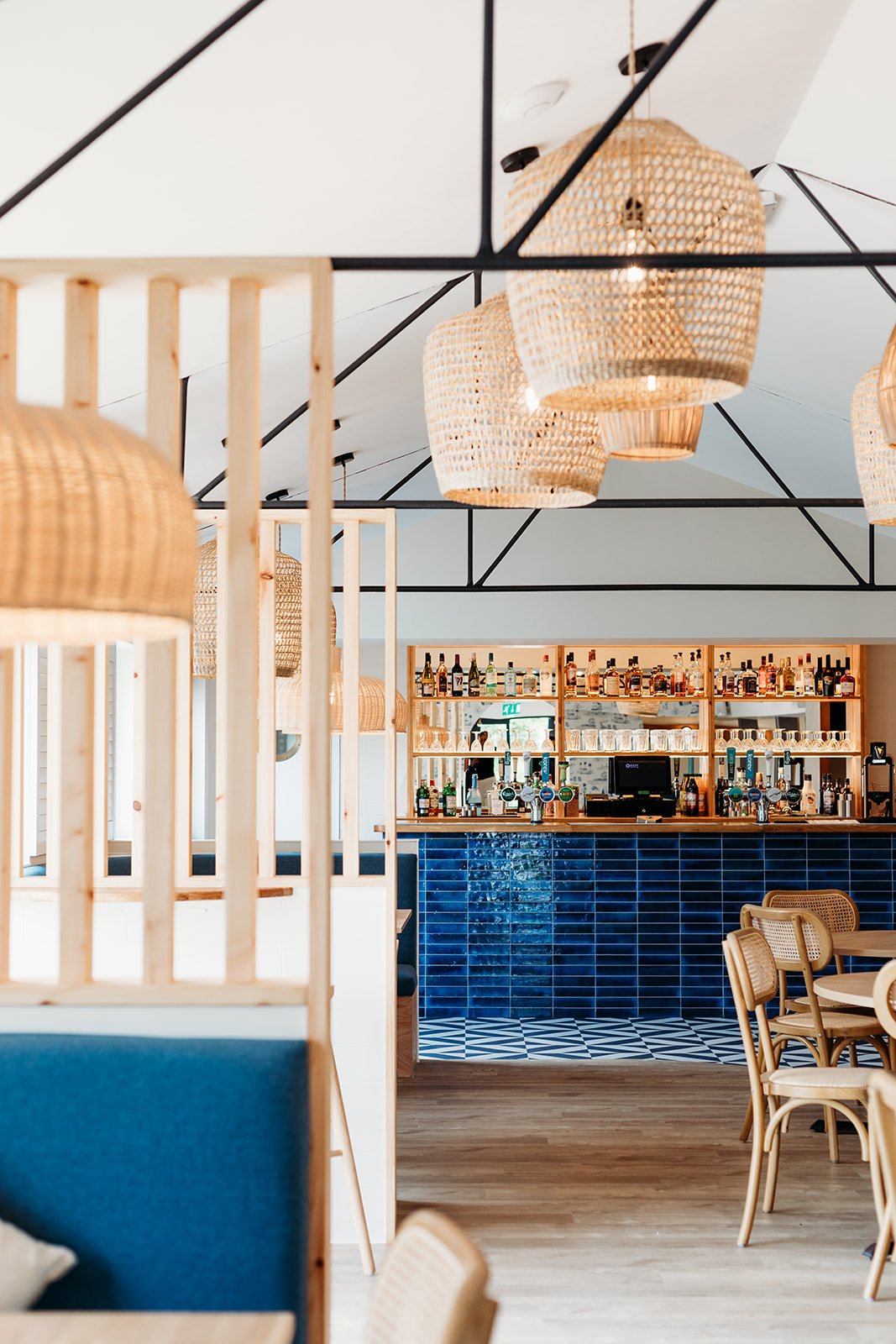 Interior of a modern restaurant or bar with rattan hanging lamps, a wooden bar counter with blue tiles, and wooden chairs and tables.