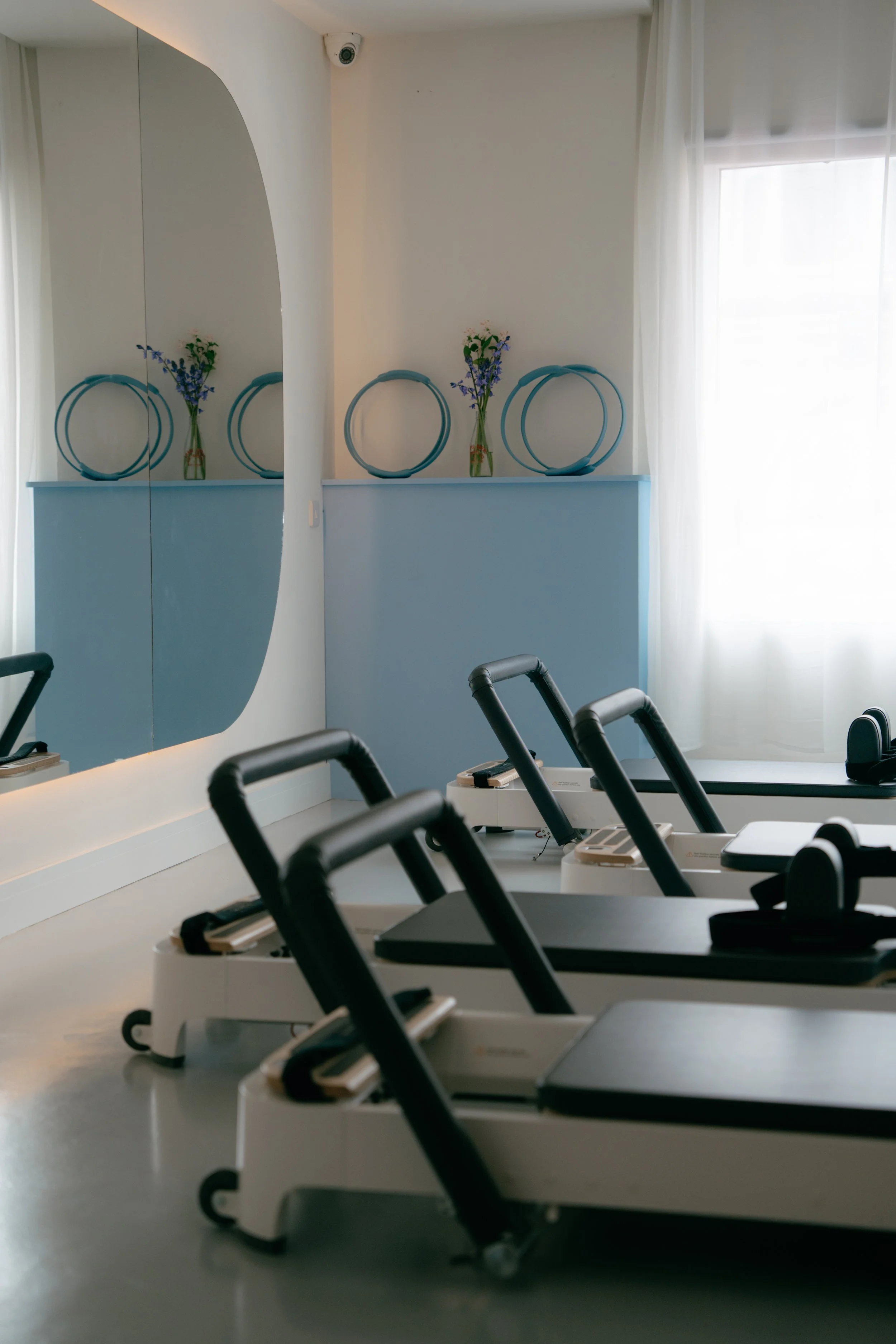 Row of white exercise or medical machines with black handles and screens, lined up on a light-colored floor in a room with bright natural light and white curtains. A blue wall with a decorative shelf holding vases with blue and white flowers is visible in the background.
