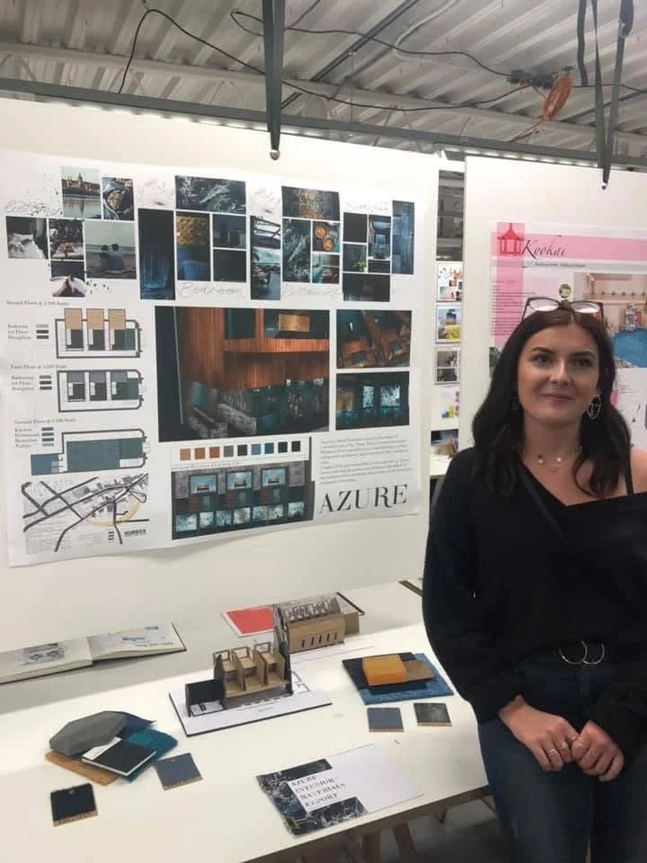 A woman standing next to an architectural presentation board at an expo or exhibition, showcasing design plans, images, and details for a project titled 'Azure' with additional design materials on the table in front.