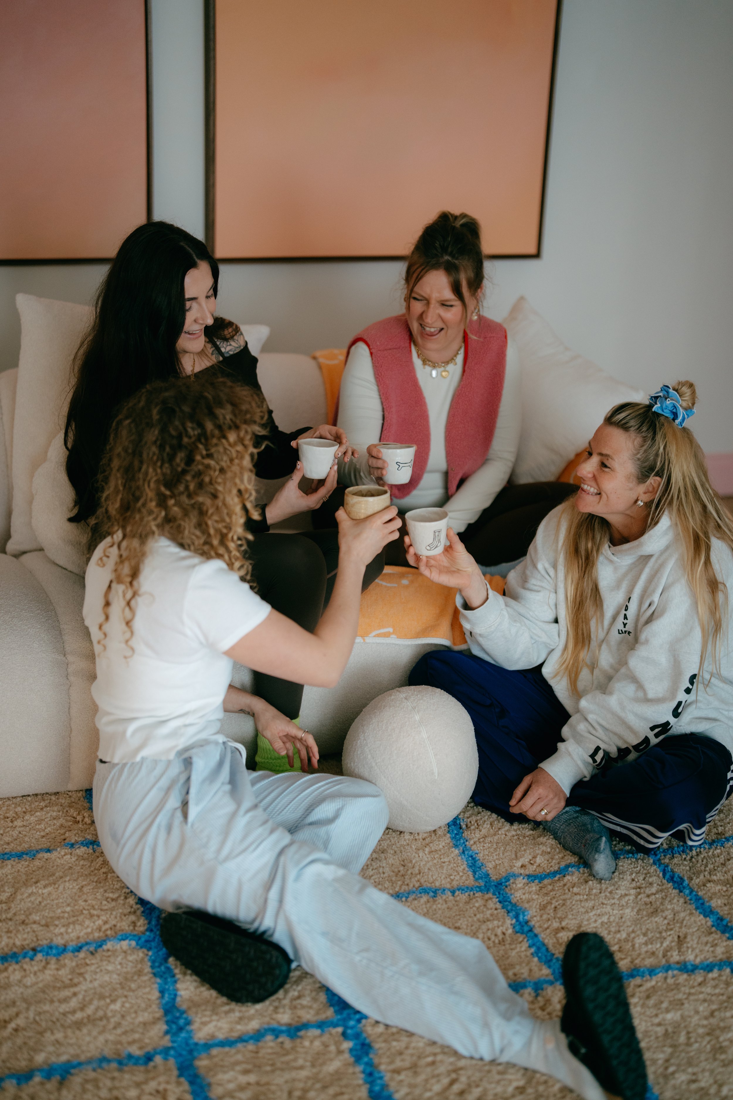Group of five women laughing and enjoying drinks together in a living room. They are sitting on and around a sofa with a multicolored rug underneath.