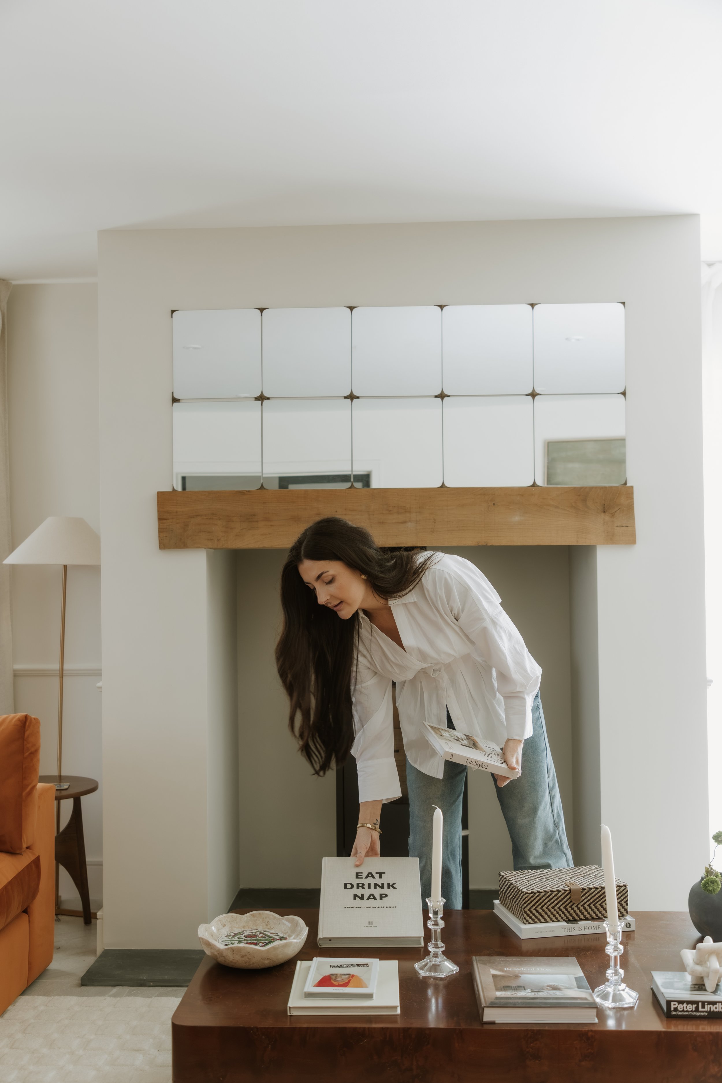 Woman in white shirt and jeans leaning over a table with decor and books, arranging candles with a living room in the background.