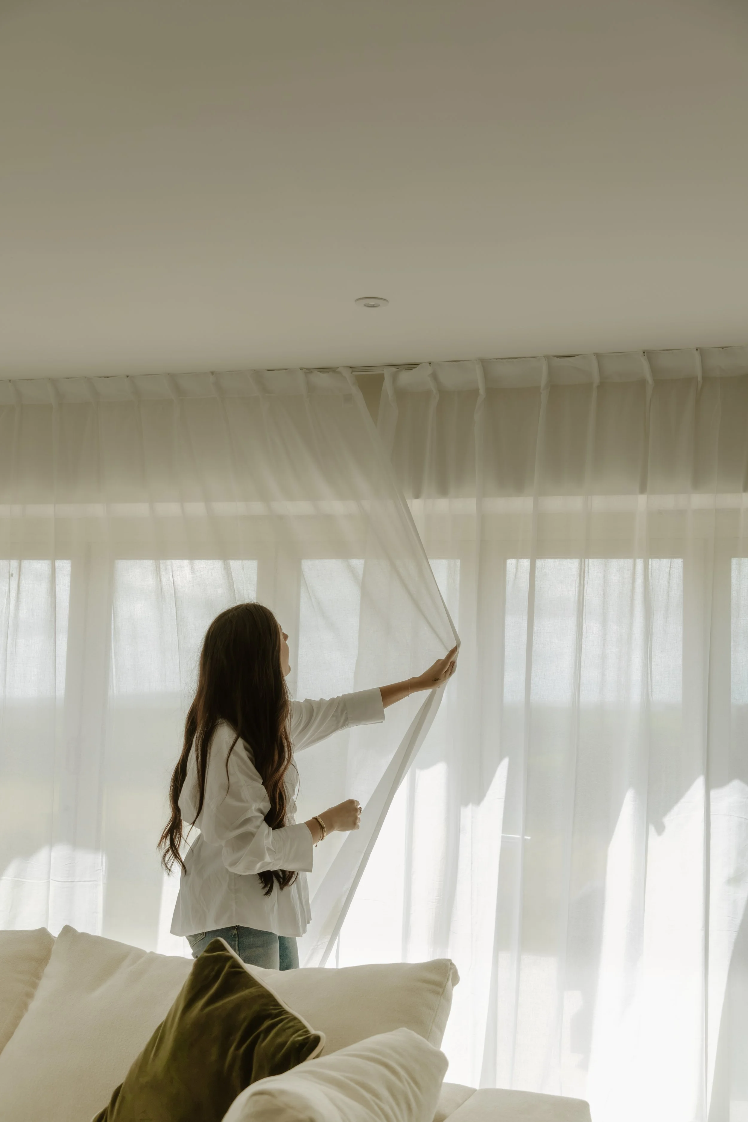 A woman with long brown hair standing near a window with sheer curtains, adjusting a curtain. A bed with brown and white pillows is visible in the foreground.