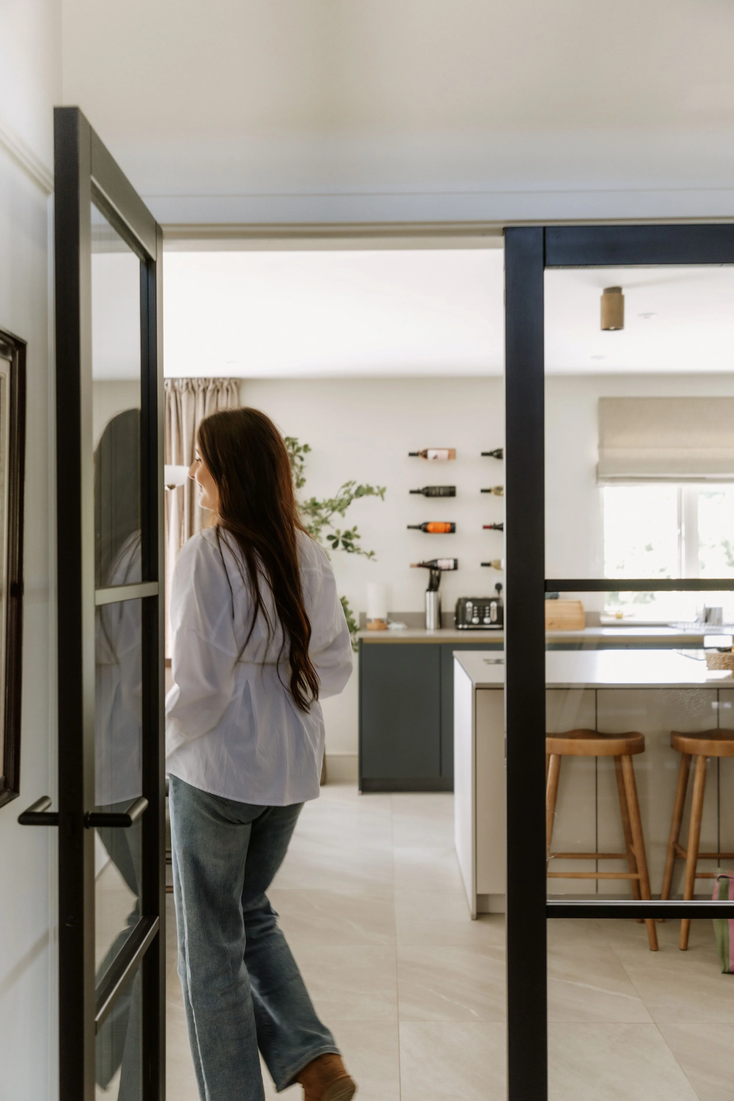 A woman with long brown hair, wearing a white shirt and jeans, standing inside a modern kitchen, looking at a mirror or glass door.