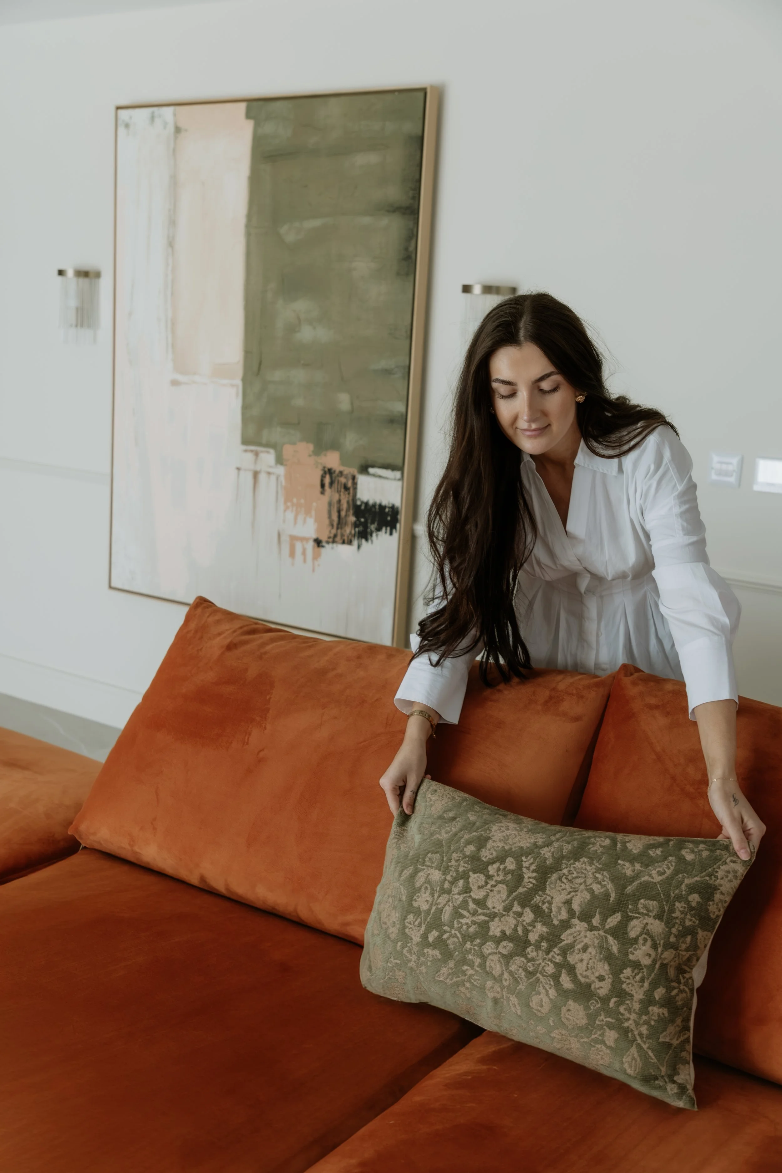 A woman with long dark hair is adjusting a decorative pillow on a burnt orange sofa in a modern, minimalist living room with abstract artwork on the wall.