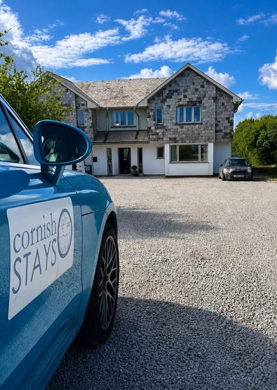 A blue car with a 'Cornish Stay' sign parked in front of a modern two-story house with a gravel driveway and a clear blue sky with some clouds.