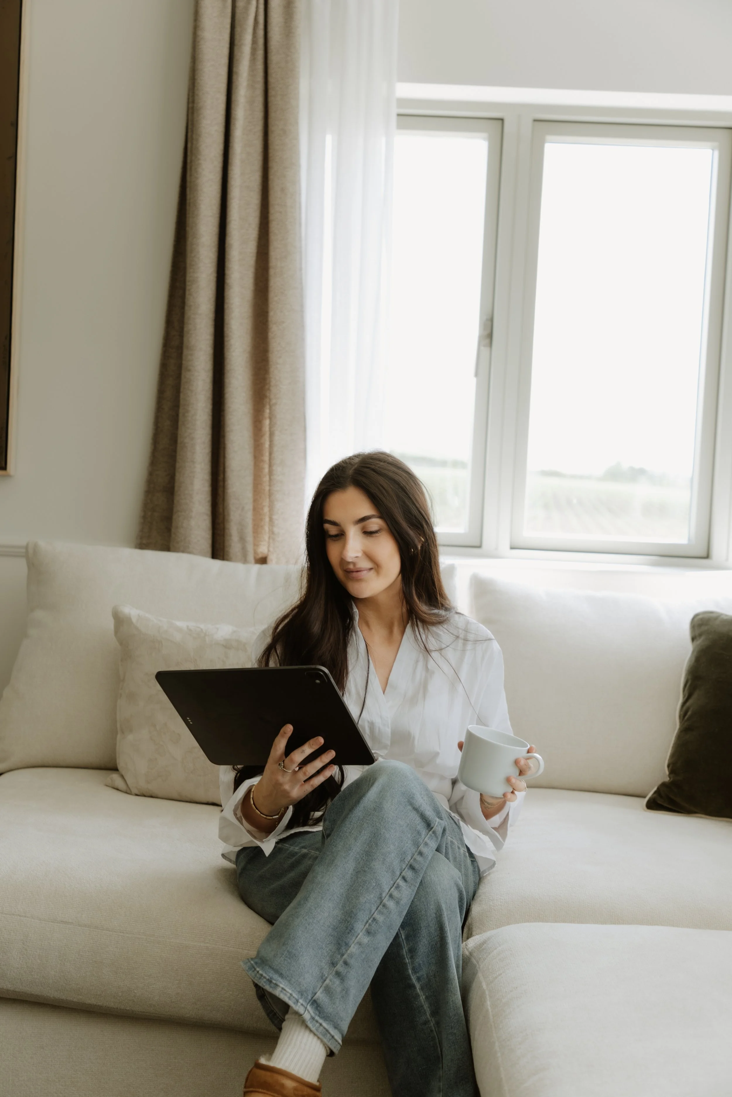 Young woman sitting on a beige couch in front of a large window with beige curtains, holding a tablet in one hand and a white mug in the other, with a relaxed and content expression.