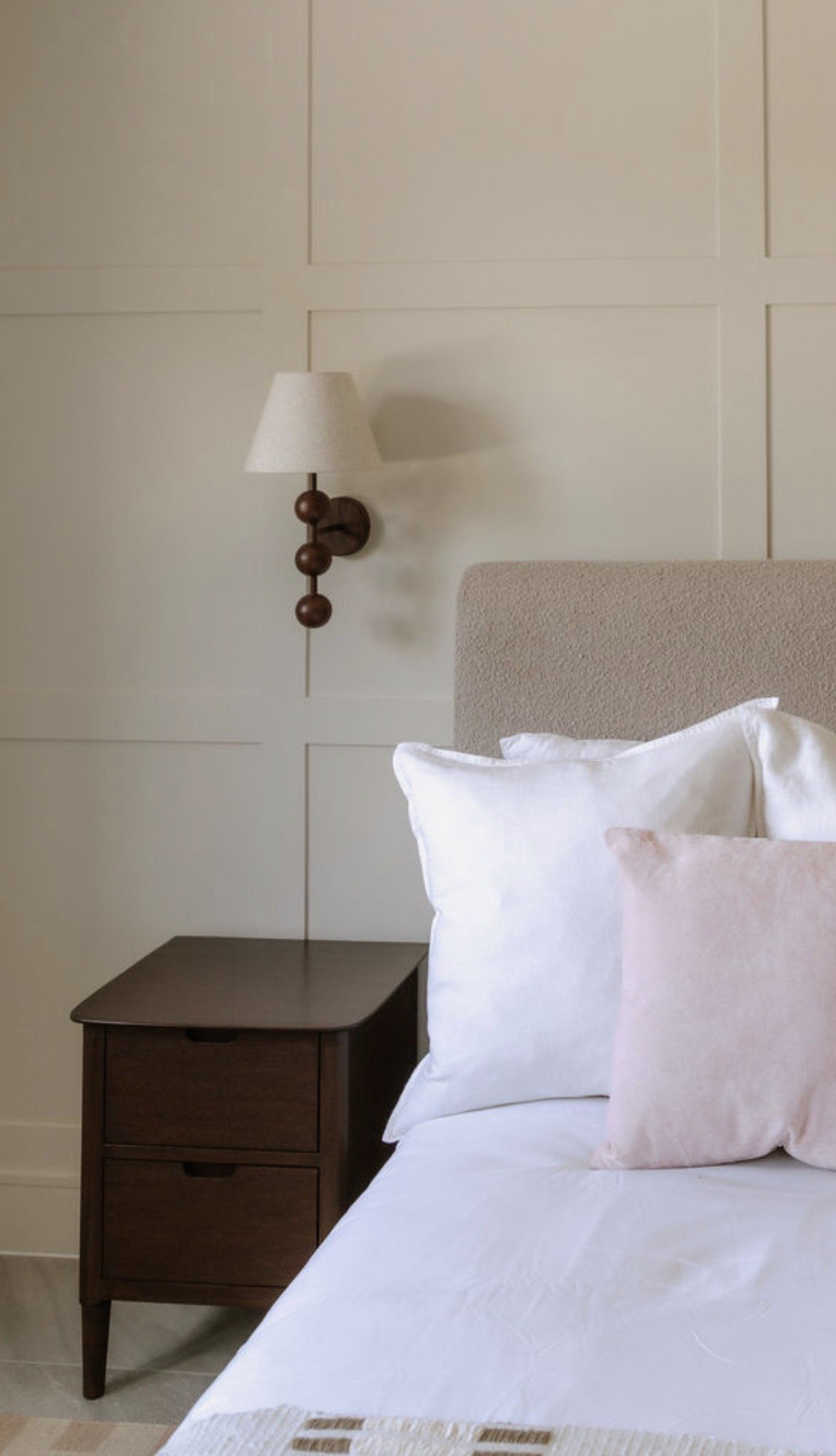 A bedroom with a bed featuring white bedding and pillows, a beige upholstered headboard, a dark wooden nightstand, and a beige wall with grid paneling. A wall-mounted sconce with three spherical wooden accents and a cone-shaped lampshade is beside the bed.