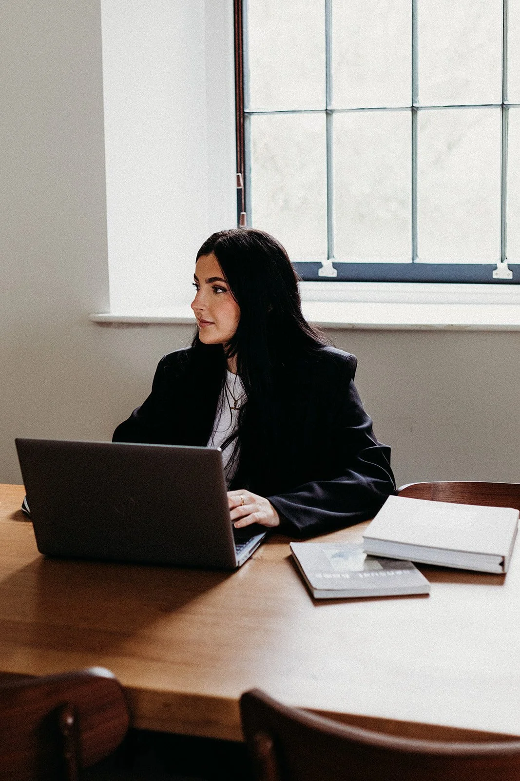 A woman with long black hair working on a laptop at a wooden table, with books and a notebook beside her, in a room with a large window.